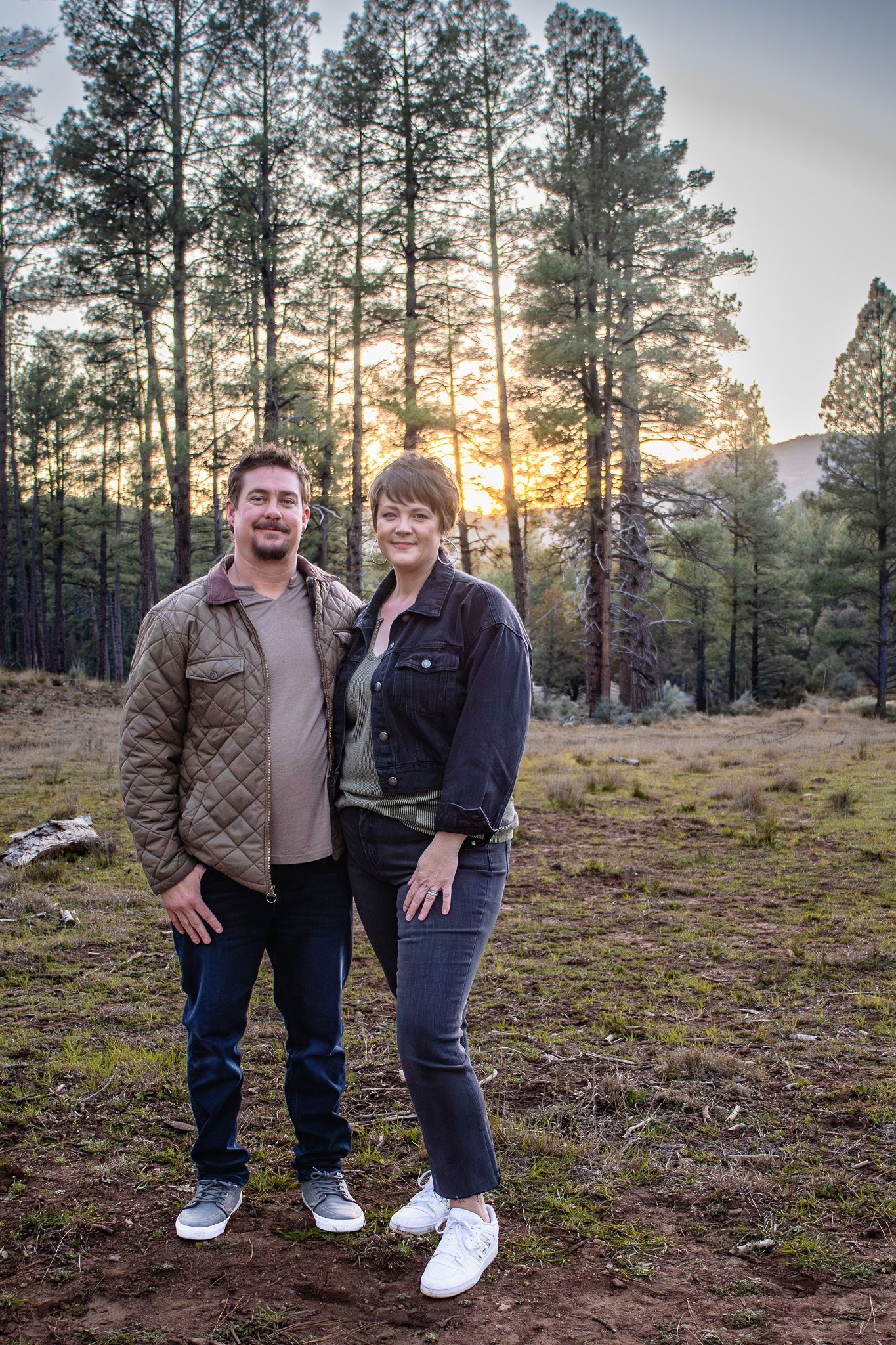 Two people standing together in a forest at sunset, with tall pine trees and mountains in the background.