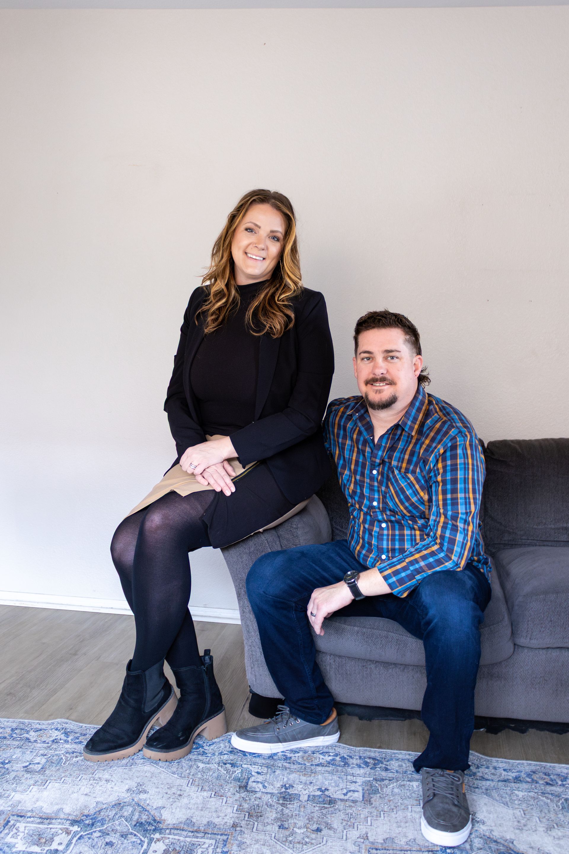 A smiling woman sits on the back of a sofa next to a man sitting on the cushions, both posing against a plain wall.