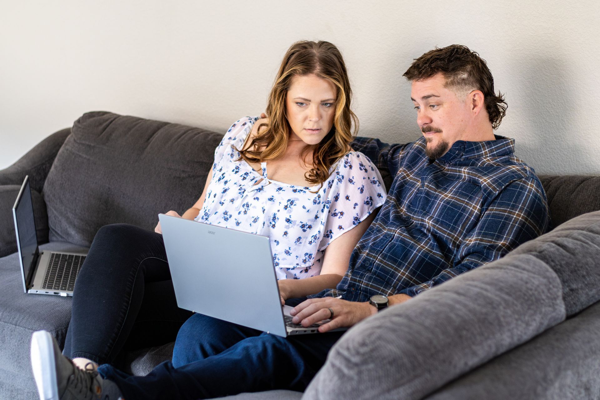 Couple on a gray couch looking at a laptop, another laptop is beside them.