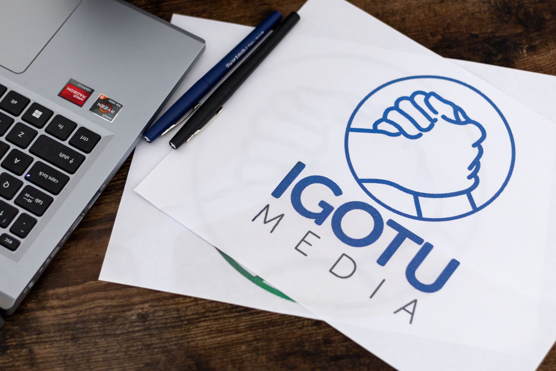 Laptop and pens on a wooden surface next to a paper featuring the blue Laptop and pens on a wooden surface next to a paper featuring the blue