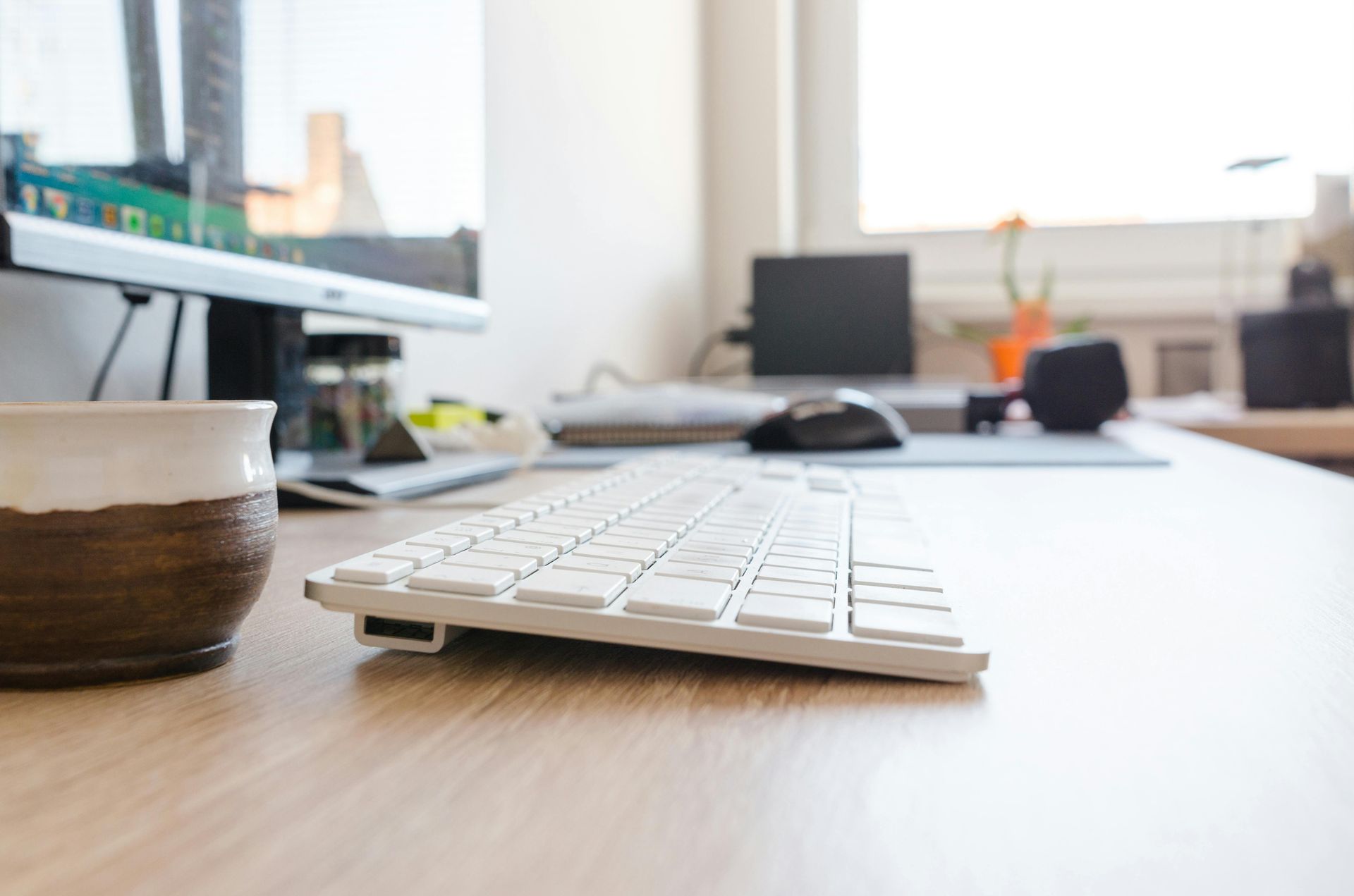 A white keyboard sits on a wooden desk next to a brown coffee mug in a bright office space.