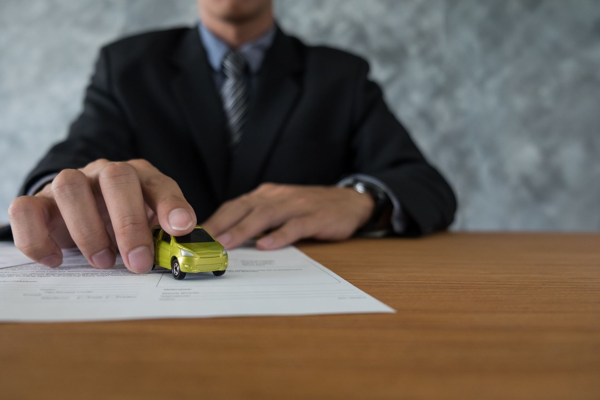 Person in a suit holding a small yellow car above paperwork on a wooden desk.