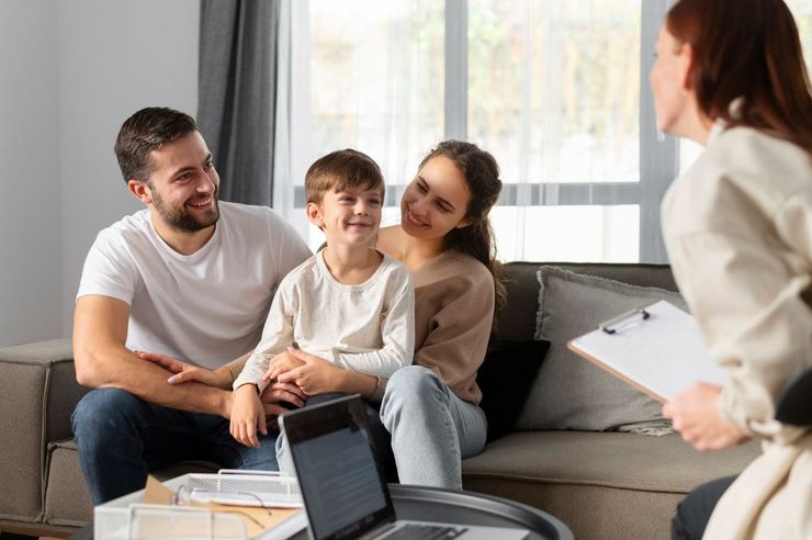 Family of three smiling, seated on a couch, consulting with a person holding a clipboard in a living room.