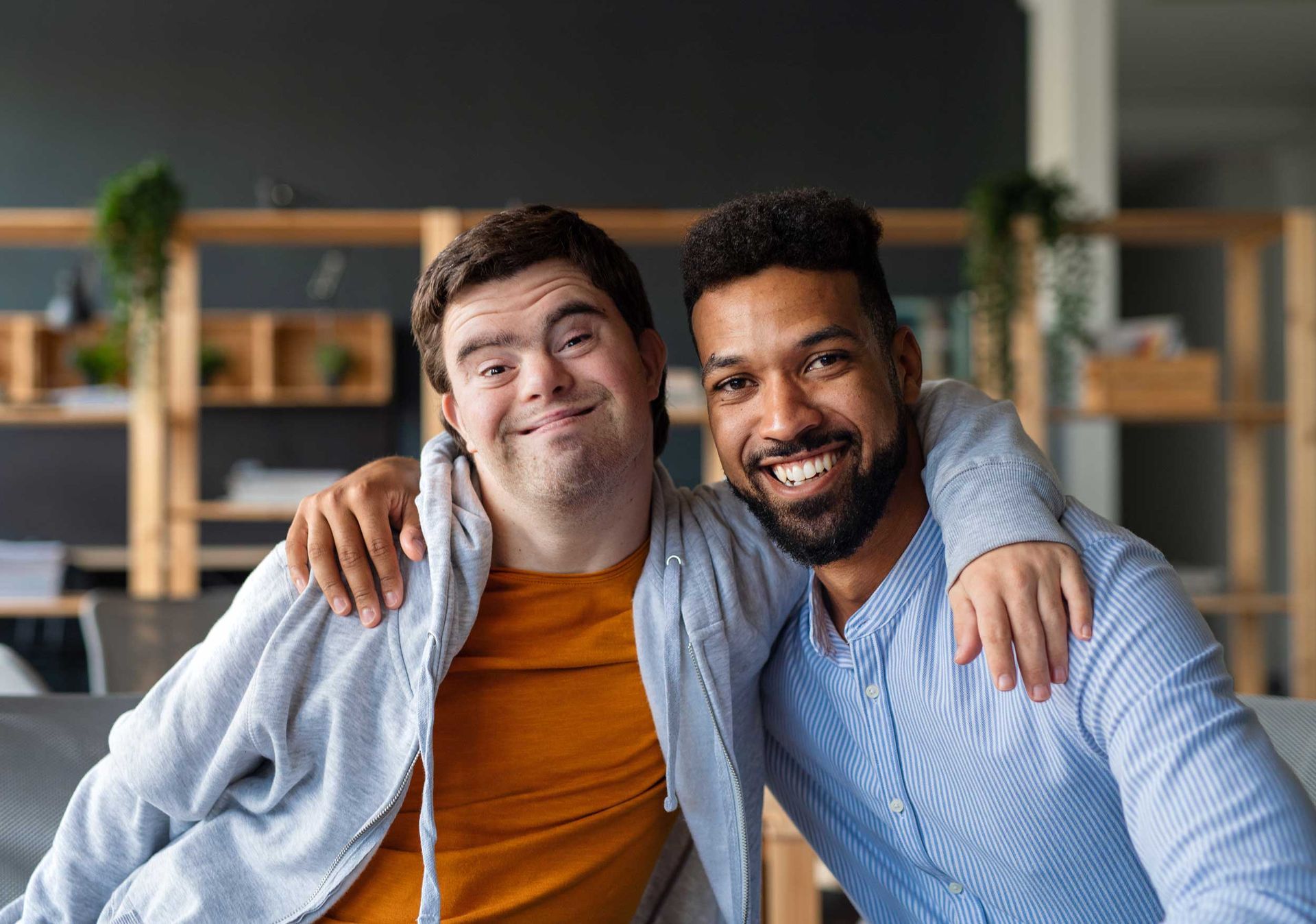 Young man with Down syndrome and his tutor with arms around him looking at the camera indoors.