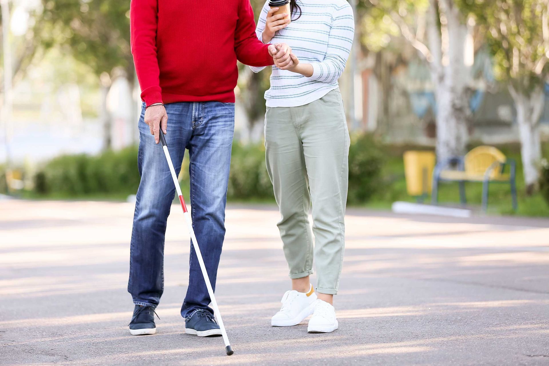 A blind man with a carer walking outdoors with his straight cane.