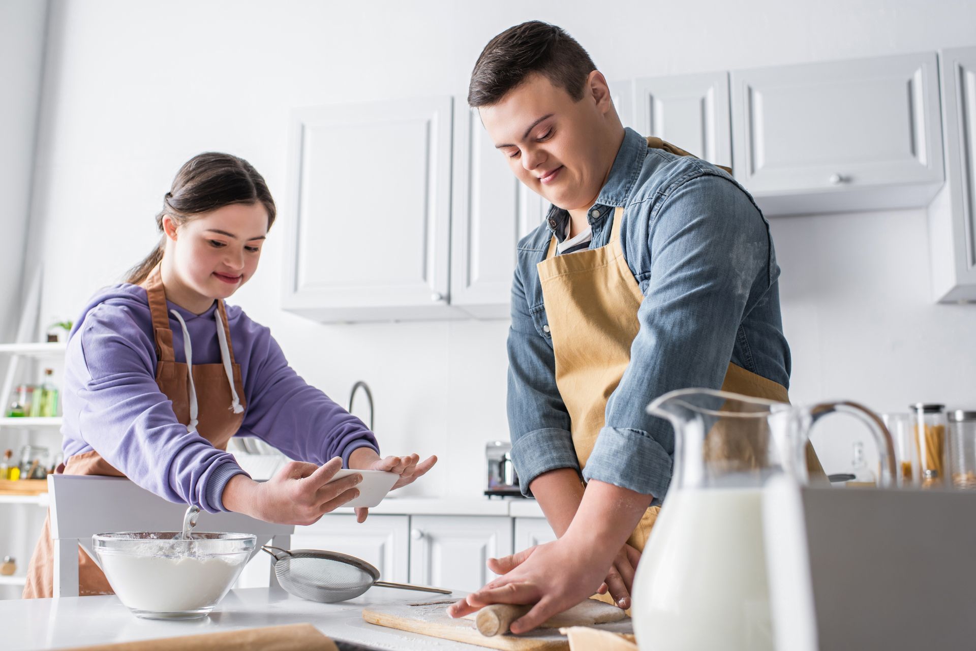 A smiling teenager with Down syndrome rolling dough near a friend taking a photo in a kitchen.