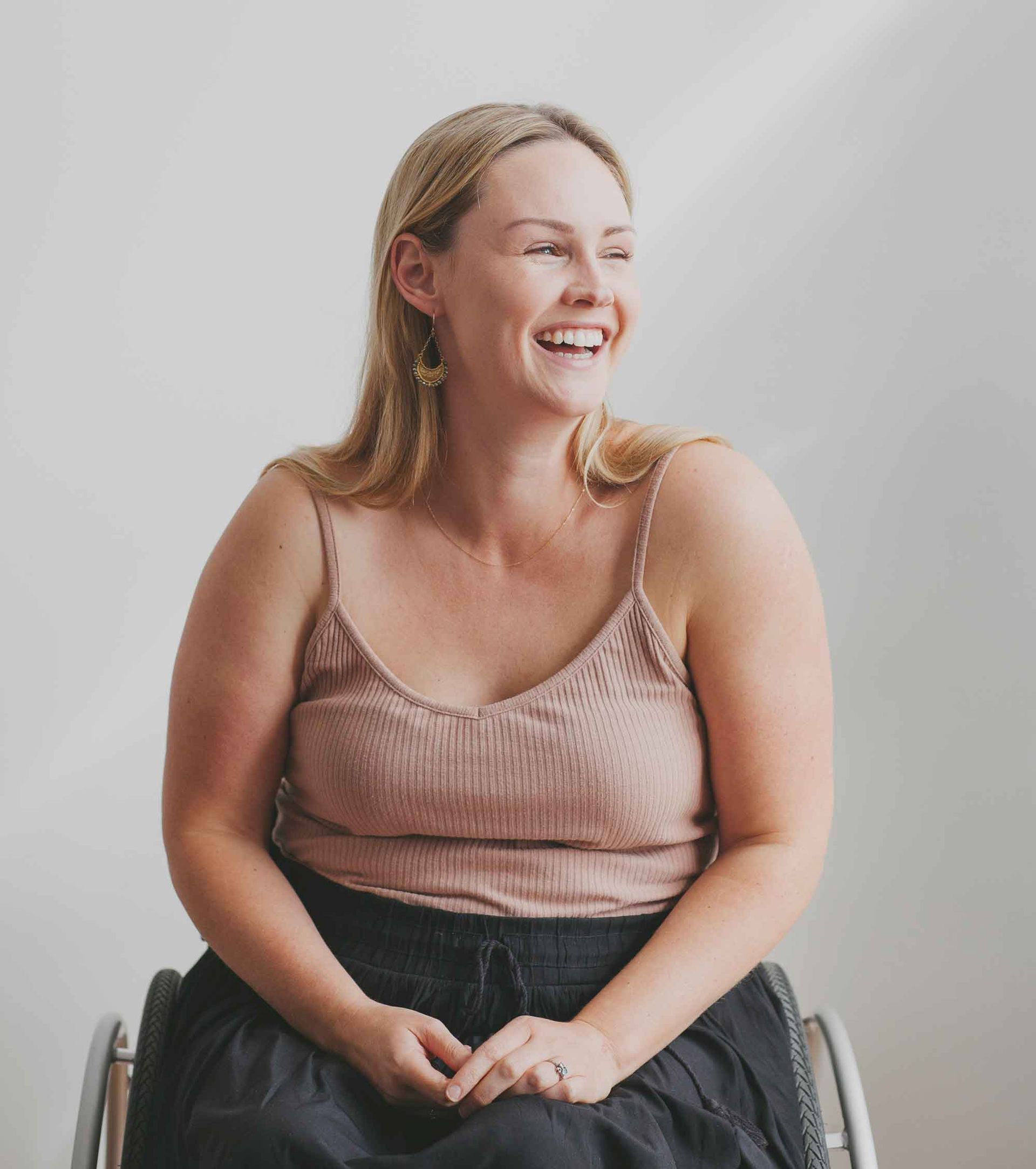 Woman in a wheelchair smiles, looking to the side. She wears a tan tank top and black skirt against a white wall. Woman in a wheelchair smiles, looking to the side. She wears a tan tank top and black skirt against a white wall.