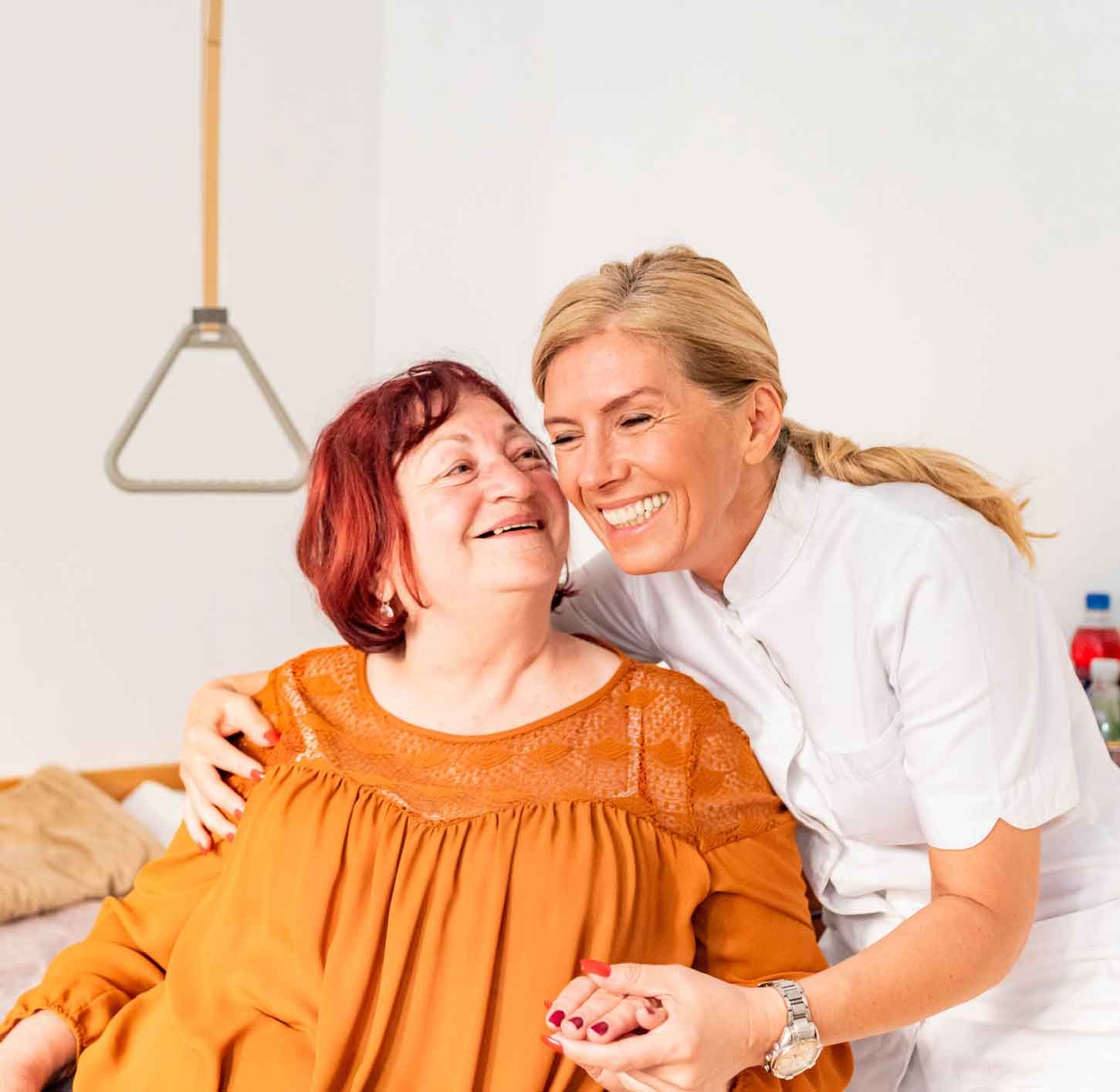A smiling caregiver embraces an older woman with red hair, offering support in a home-care setting.