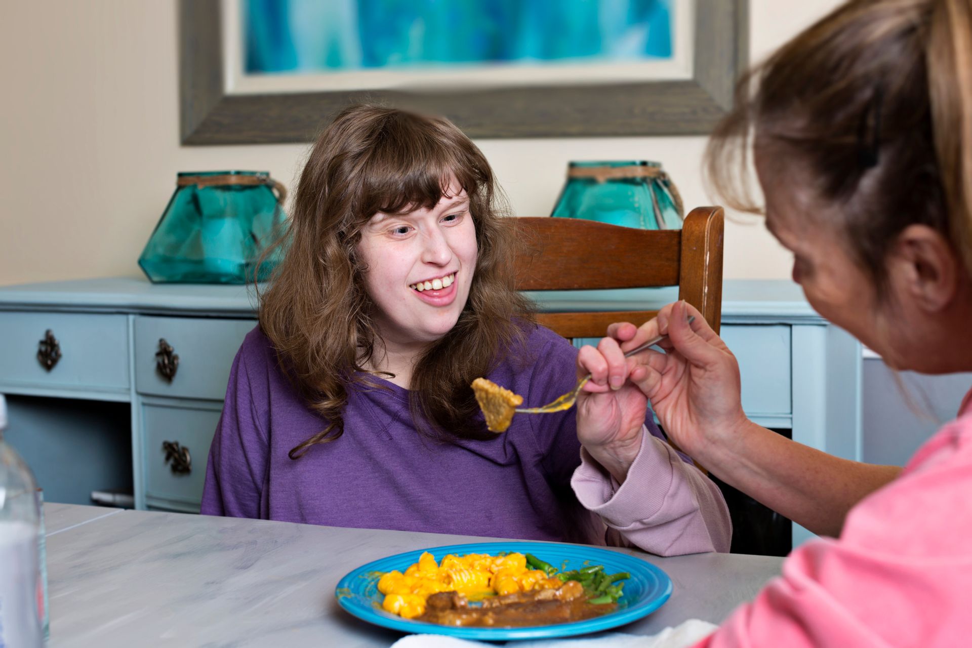 Woman with Down syndrome being fed by another woman at a table; food on blue plate, light blue furniture.