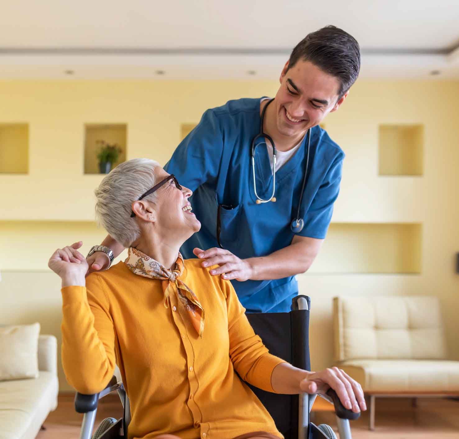 A smiling healthcare worker assists an elderly woman in a wheelchair indoors. Both are looking at each other, the woman in an orange sweater, and the worker in blue scrubs.