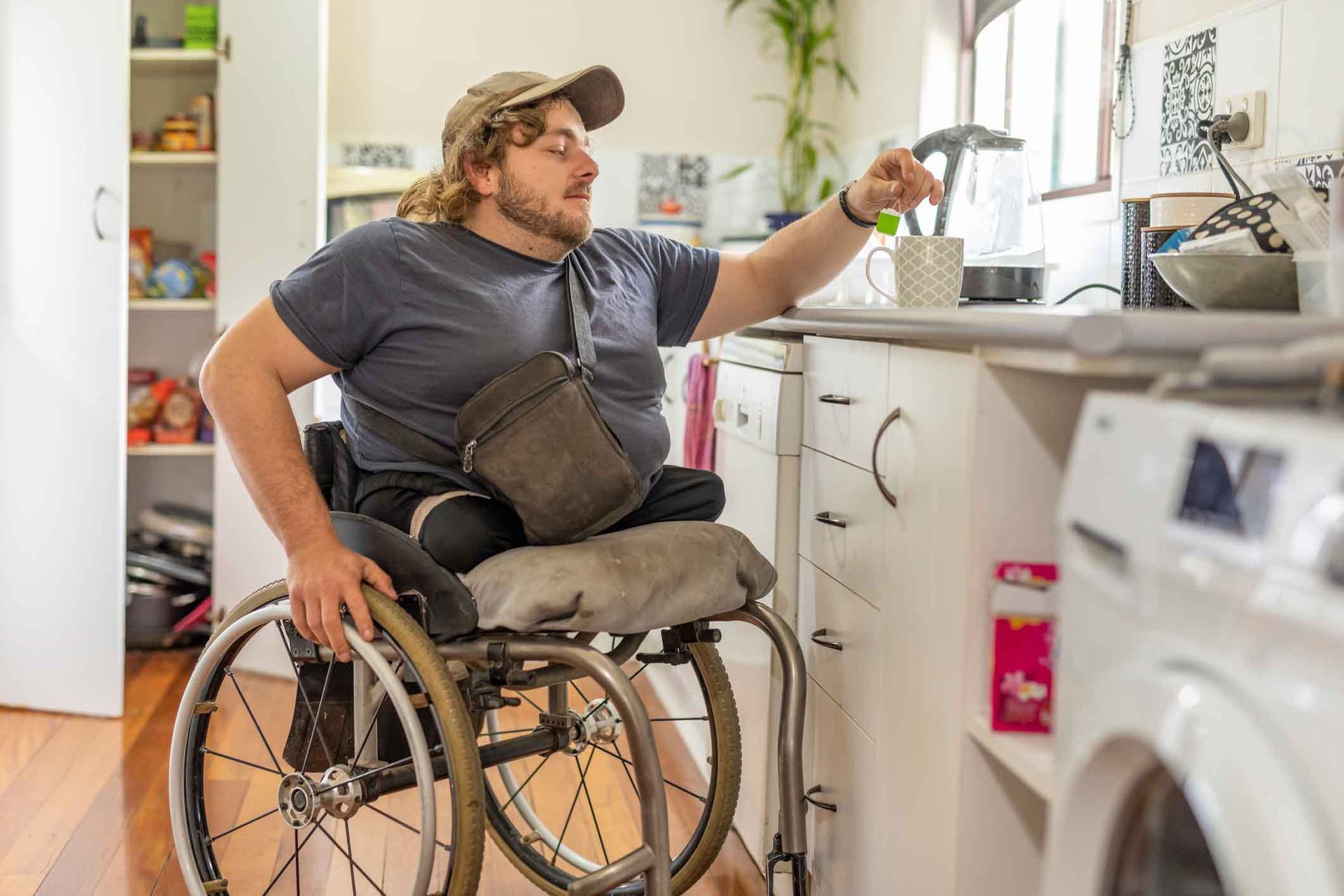 A man in a wheelchair reaches toward a window in a kitchen. He wears a cap and blue shirt. White cabinets and appliances are visible. A man in a wheelchair reaches toward a window in a kitchen. He wears a cap and blue shirt. White cabinets and appliances are visible.