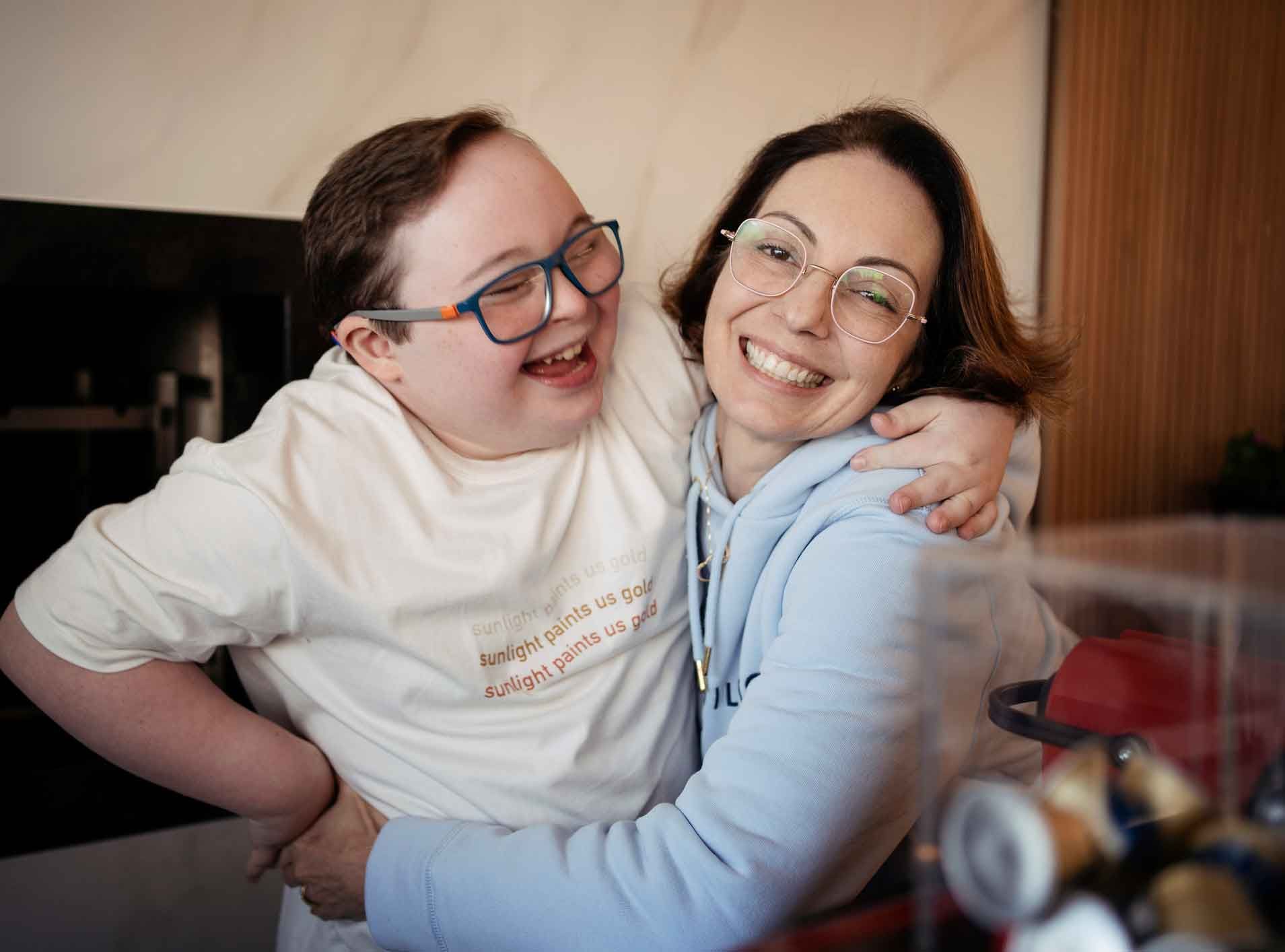 A woman and a young man with Down syndrome embrace and smile joyfully. They are indoors, with the man wearing glasses and a white t-shirt.