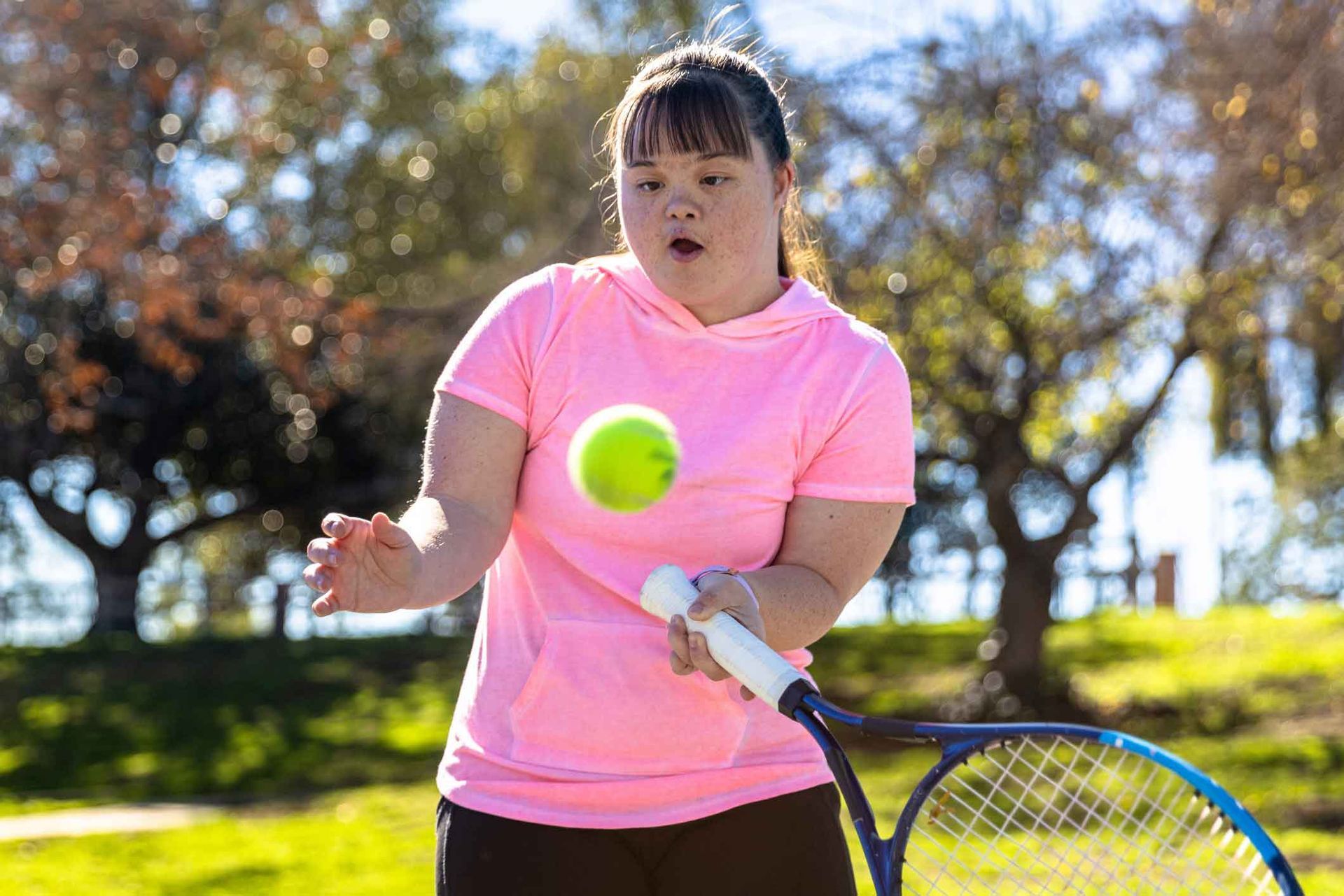A young woman with Down syndrome in a pink shirt plays tennis outdoors. She focuses on the ball, ready to hit it with her racquet.