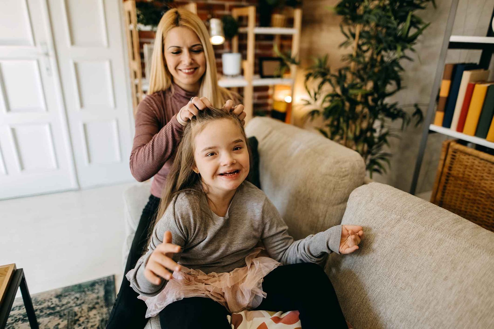 Woman braiding a young girl's hair on a sofa in a living room. Both are smiling; the girl holds a toy.