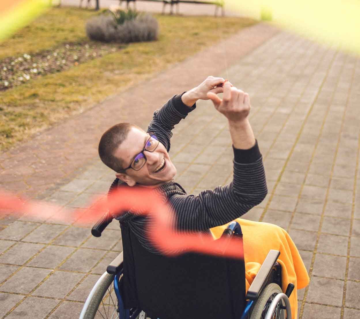 Smiling person in a wheelchair outdoors, raising their arms. They wear glasses and a dark jacket, orange blanket draped on chair.