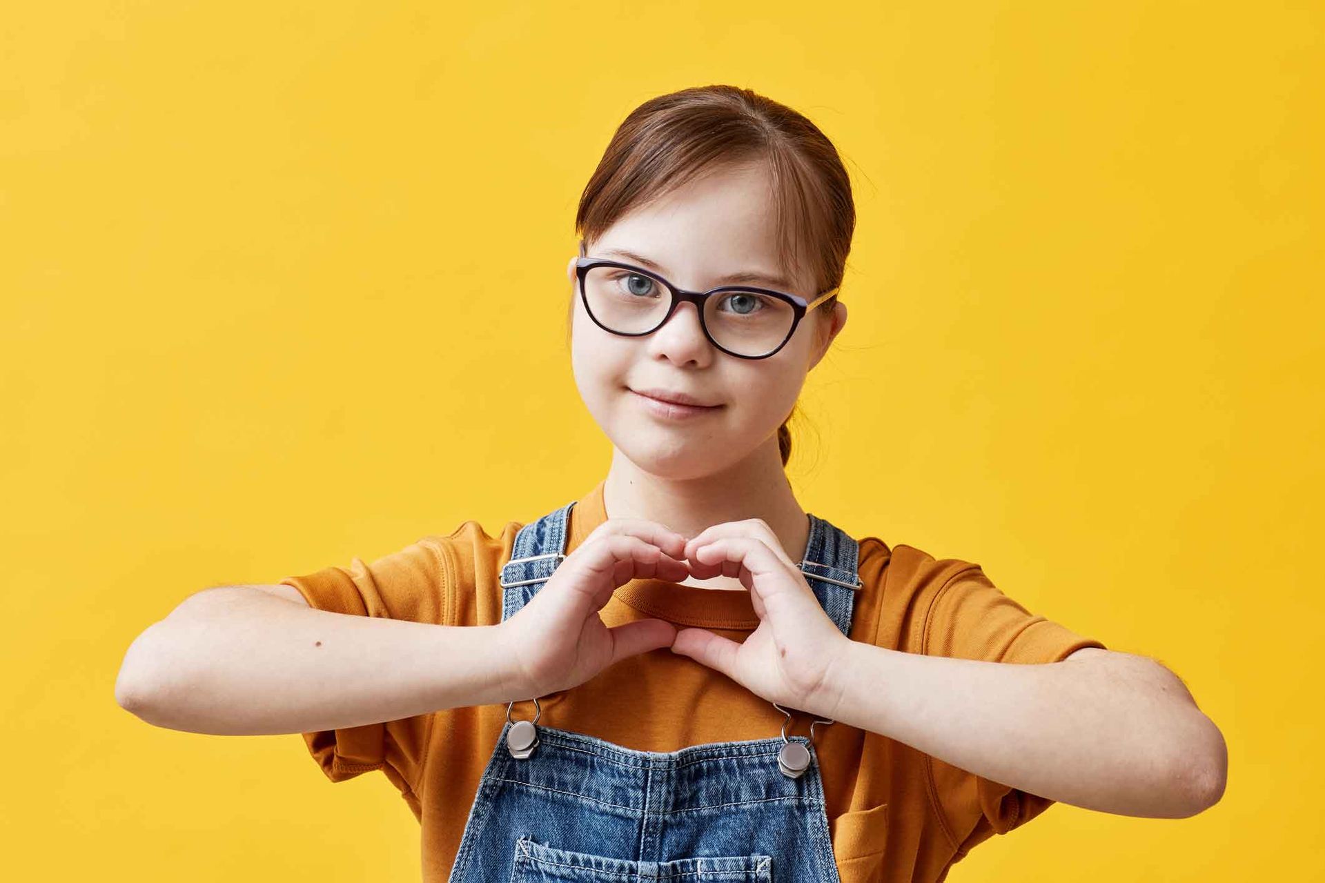 Girl with Down syndrome wearing glasses, making a heart shape with her hands, against a yellow background.