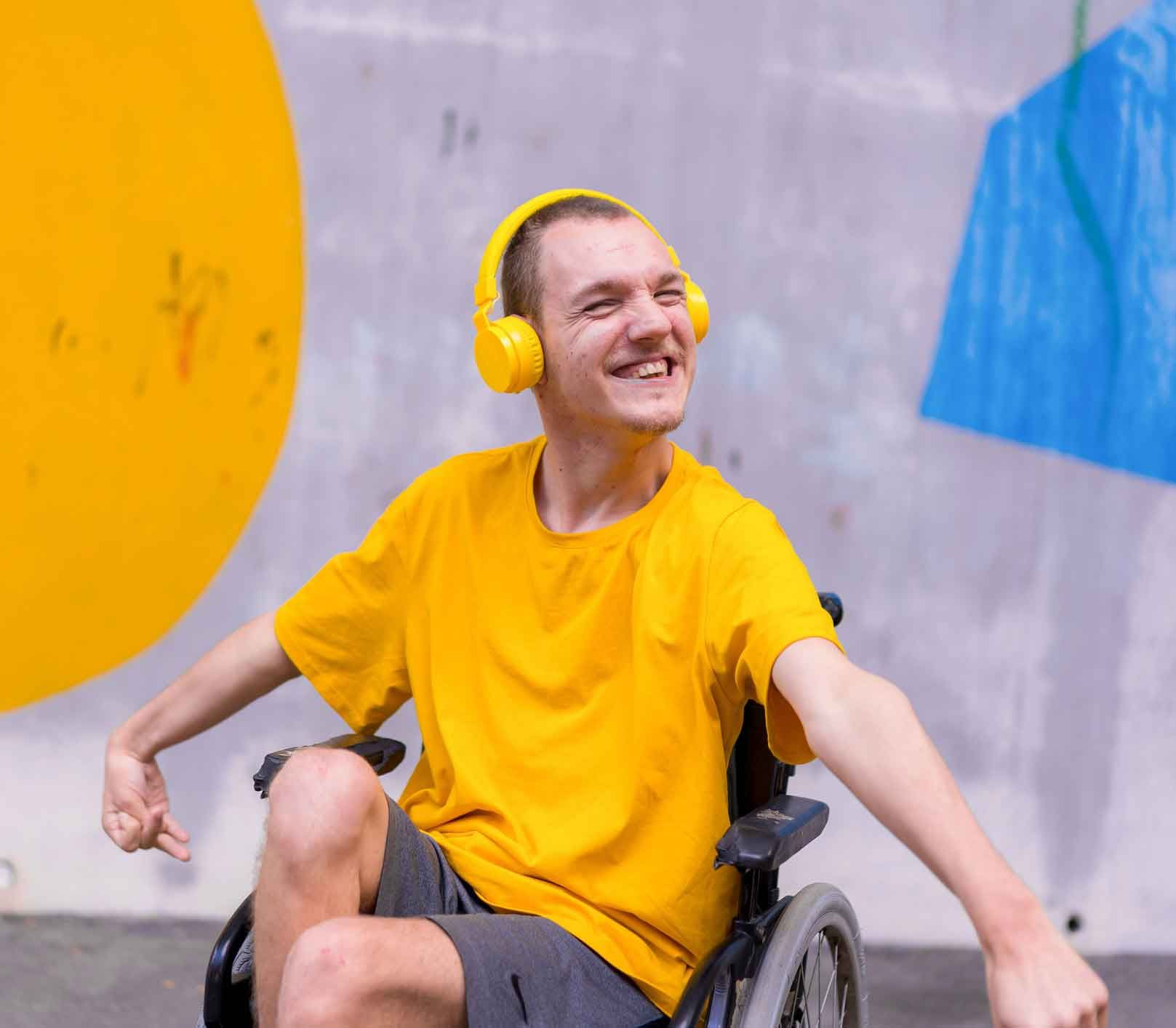 Person in a wheelchair wearing yellow headphones and shirt smiles broadly, arms outstretched, in front of colorful climbing wall.