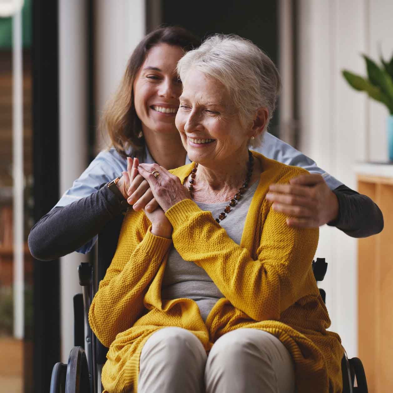 A smiling caregiver embraces an elderly woman in a wheelchair, both indoors. The woman wears a yellow sweater, and the caregiver has on a blue uniform. A smiling caregiver embraces an elderly woman in a wheelchair, both indoors. The woman wears a yellow sweater, and the caregiver has on a blue uniform.