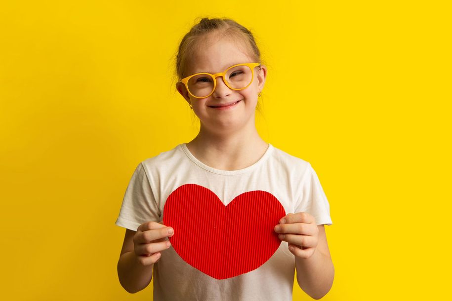 Girl with Down syndrome wearing glasses, holding a red heart, smiling against a yellow backdrop. Girl with Down syndrome wearing glasses, holding a red heart, smiling against a yellow backdrop.