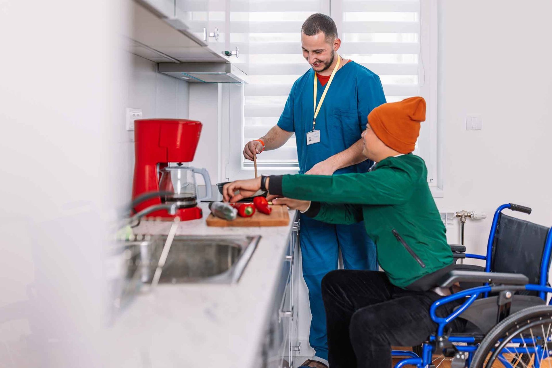 A person in a wheelchair cuts vegetables with assistance from a caregiver in a kitchen. The caregiver is wearing blue scrubs and the person in the wheelchair is wearing a green jacket and orange beanie.