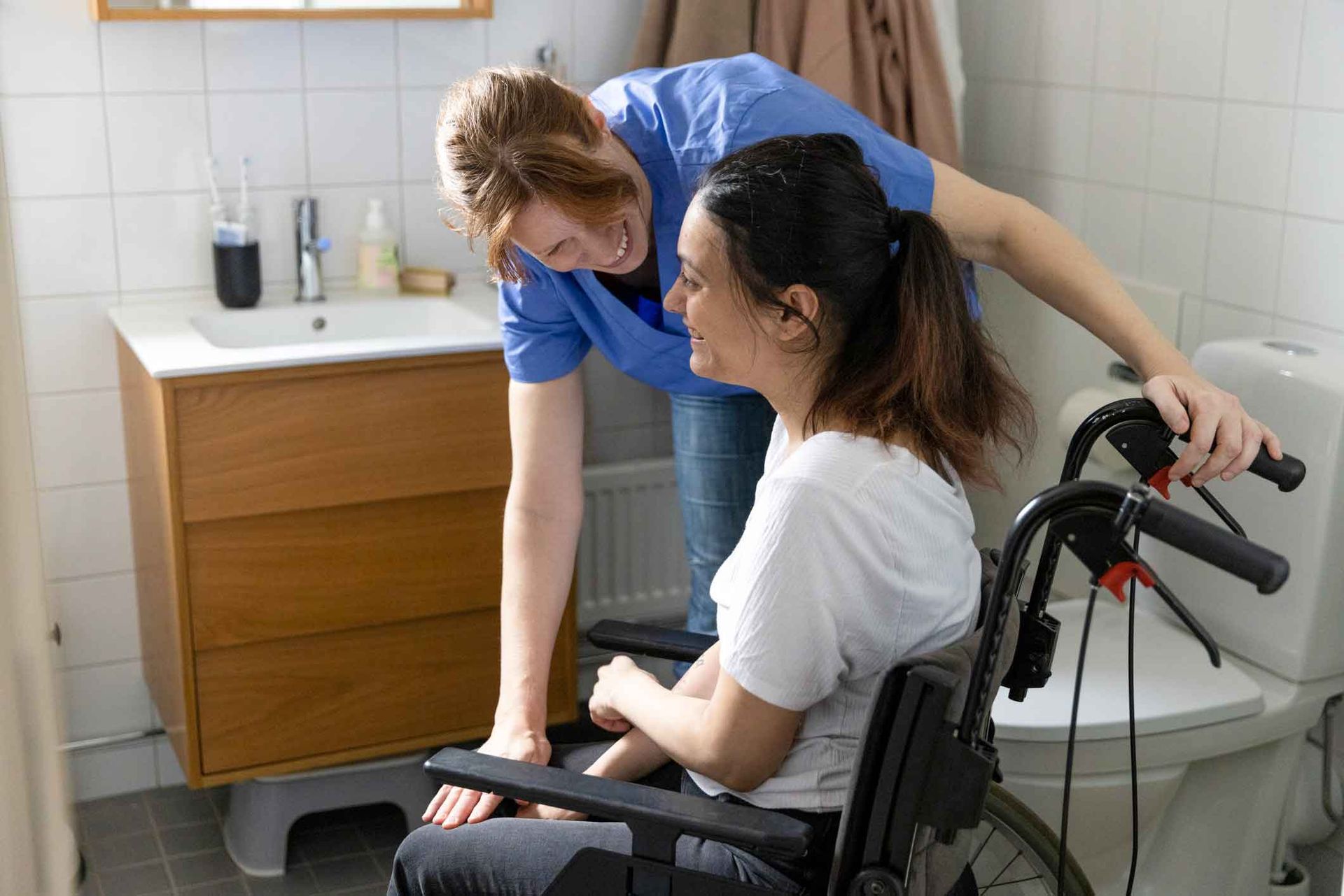 A healthcare worker assists a woman in a wheelchair near a toilet in a bathroom. The worker wears blue; the woman, a white shirt.