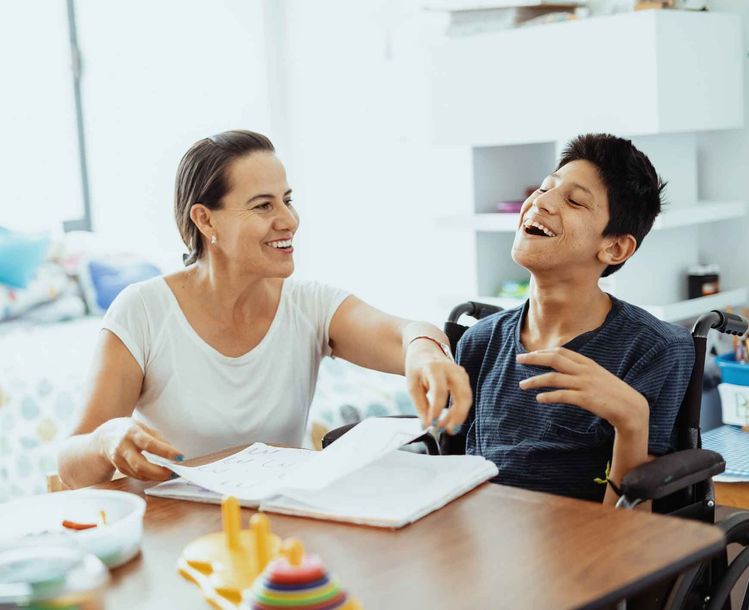 Woman and teen in a wheelchair smile at a table, looking at a notebook. Bright room, likely at home. Woman and teen in a wheelchair smile at a table, looking at a notebook. Bright room, likely at home.