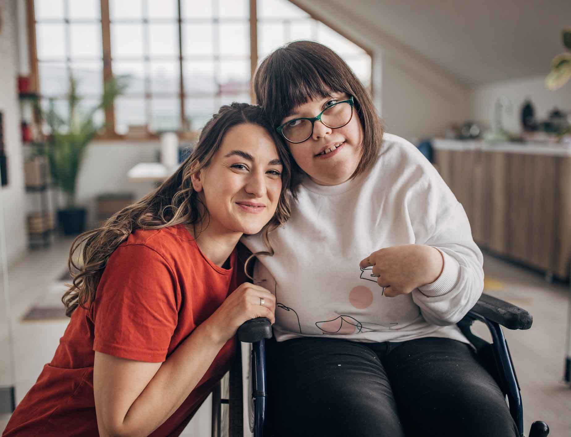 Two smiling women, one in a wheelchair, pose together indoors. The woman on the left has long brown hair, and the woman on the right wears glasses and a white sweater.