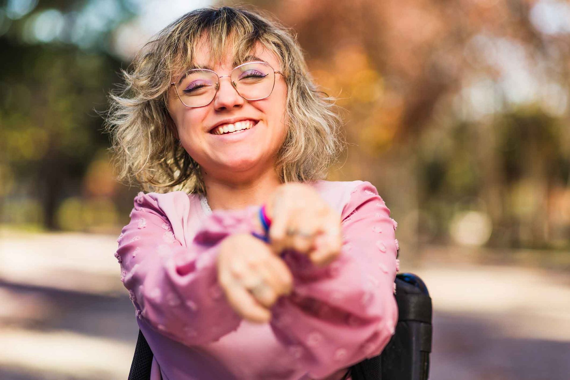 Woman in a wheelchair smiles, holding up hands. She has short blonde hair, glasses, and a pink shirt, in a park. Woman in a wheelchair smiles, holding up hands. She has short blonde hair, glasses, and a pink shirt, in a park.