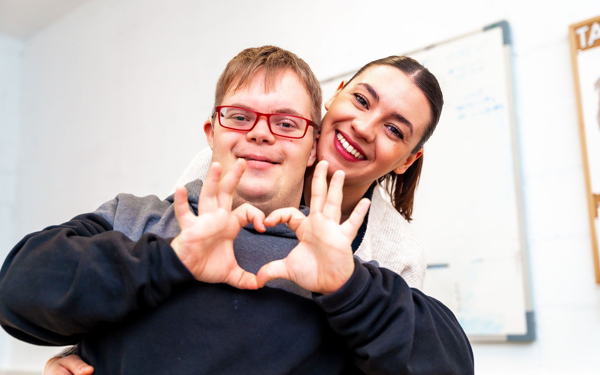 A young man with Down syndrome and a woman make a heart shape with their hands, smiling at the camera.