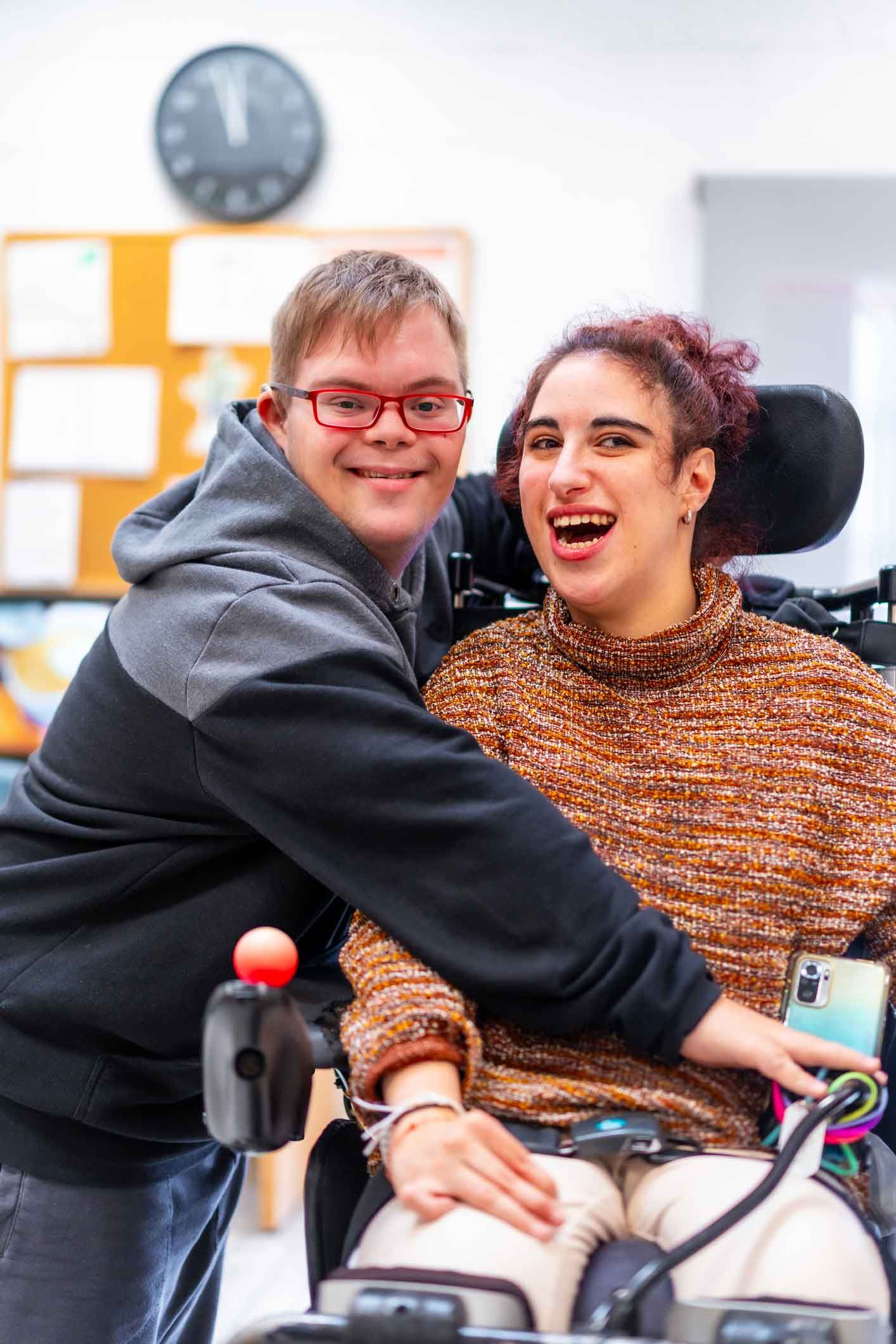 Man with Down syndrome embraces a woman in a wheelchair; both are smiling and laughing in a brightly lit room.