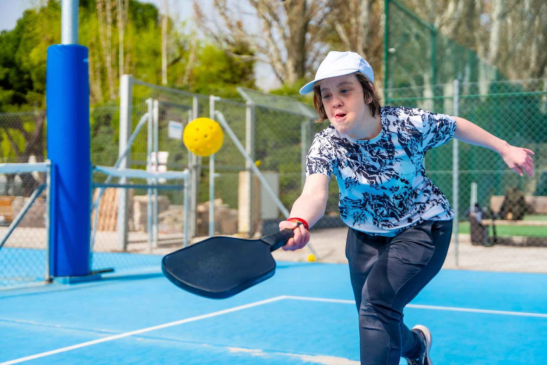 Person in white hat playing pickleball on blue court, swinging paddle at yellow ball.