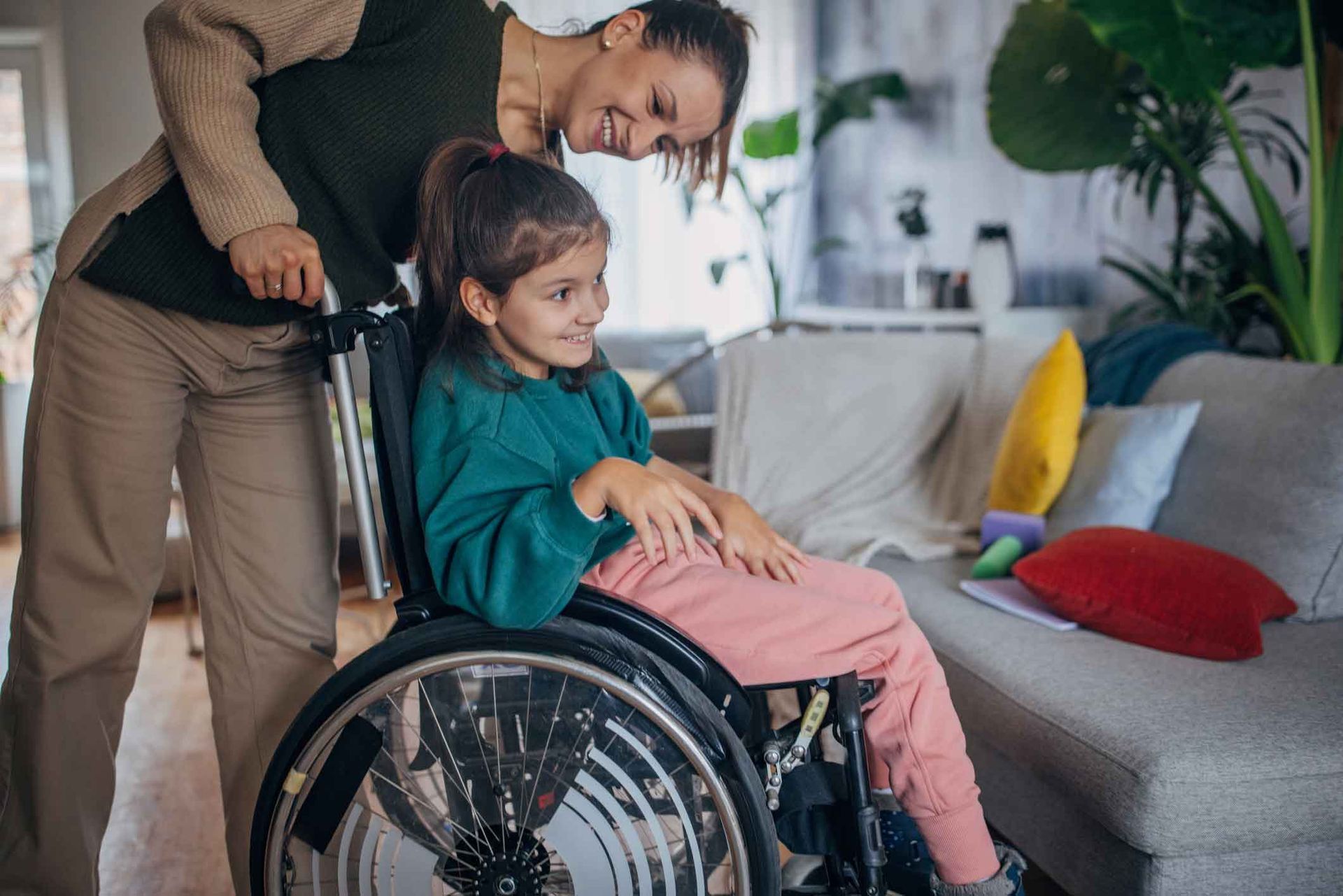 A woman smiles at a young girl in a wheelchair. The girl is sitting indoors, looking to the side, as the woman pushes her.