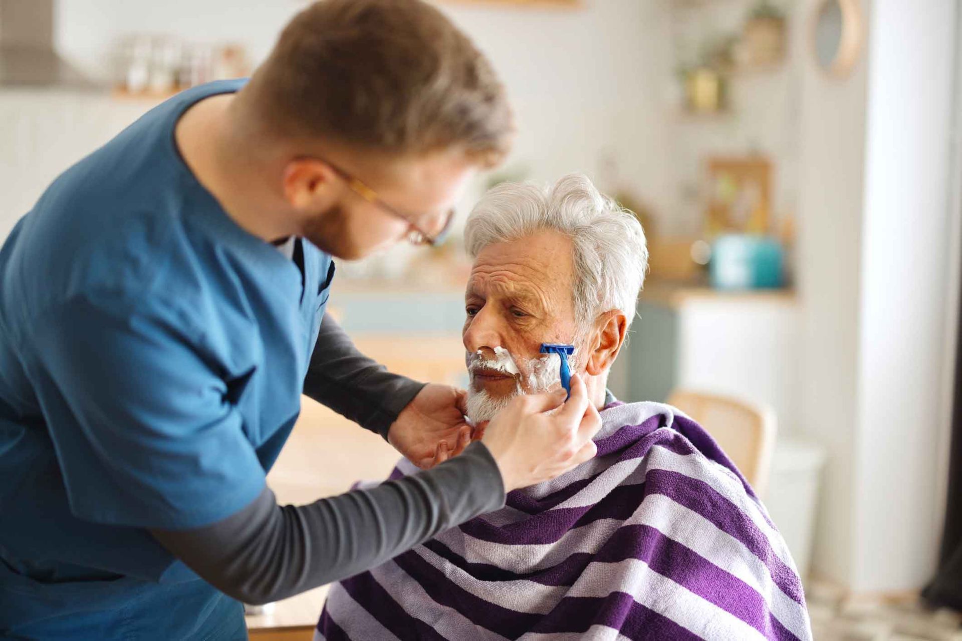 Caregiver shaves an elderly man's face with a razor in a home setting. The man is seated and covered with a striped towel.
