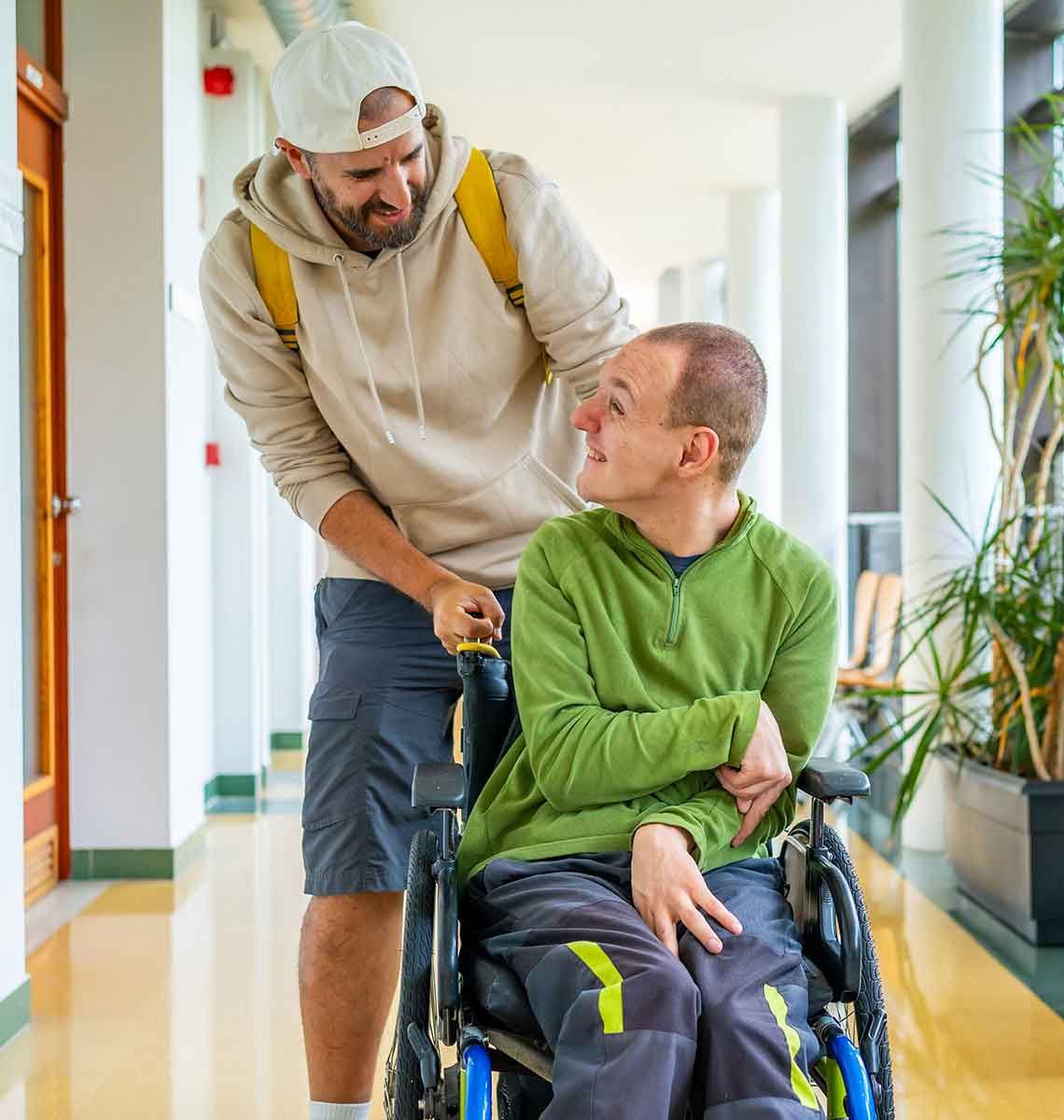 Man pushing a wheelchair with another man inside down a brightly lit hallway. Both are smiling and looking at each other.
