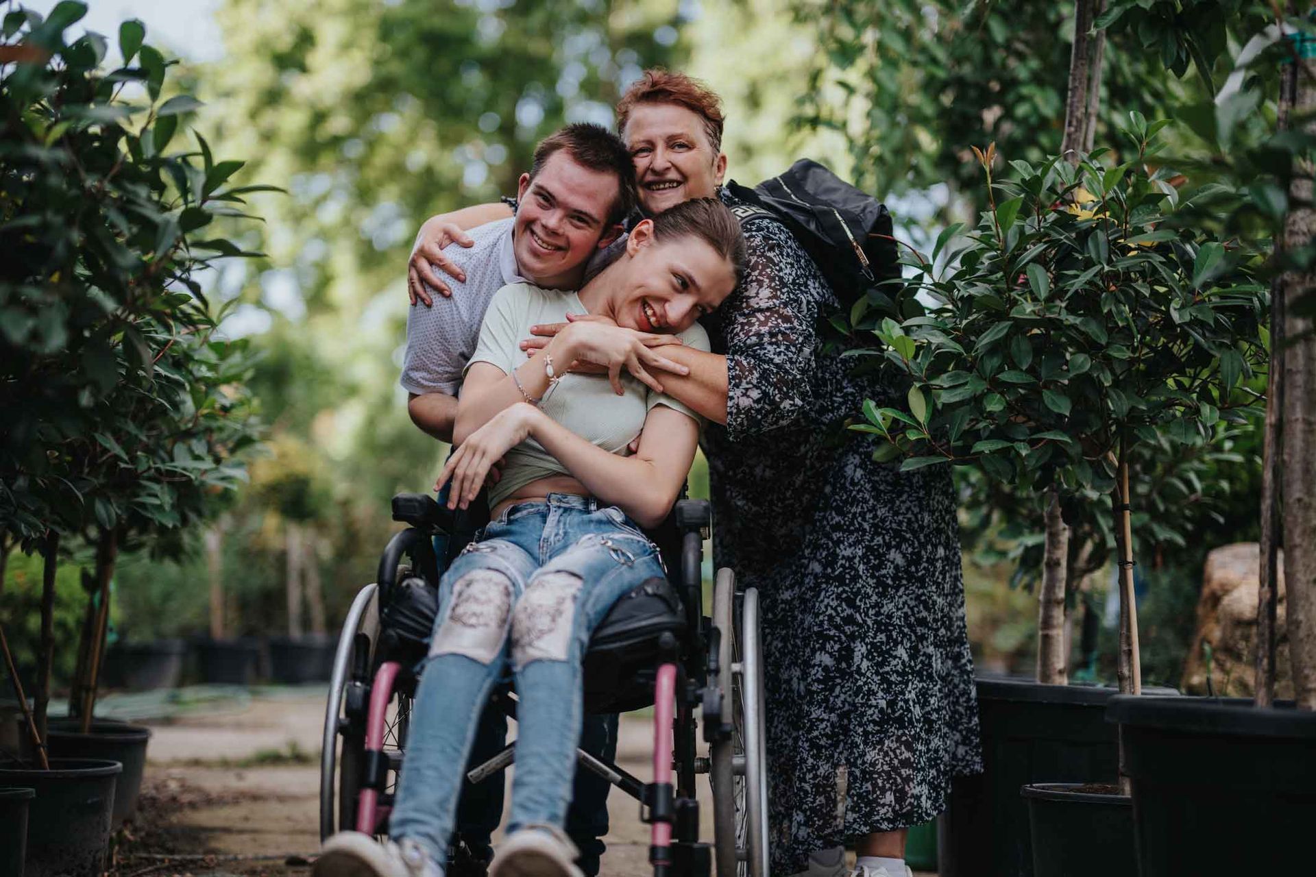 Three smiling people hugging: a woman in a wheelchair, a man, and another woman, in a plant nursery.
