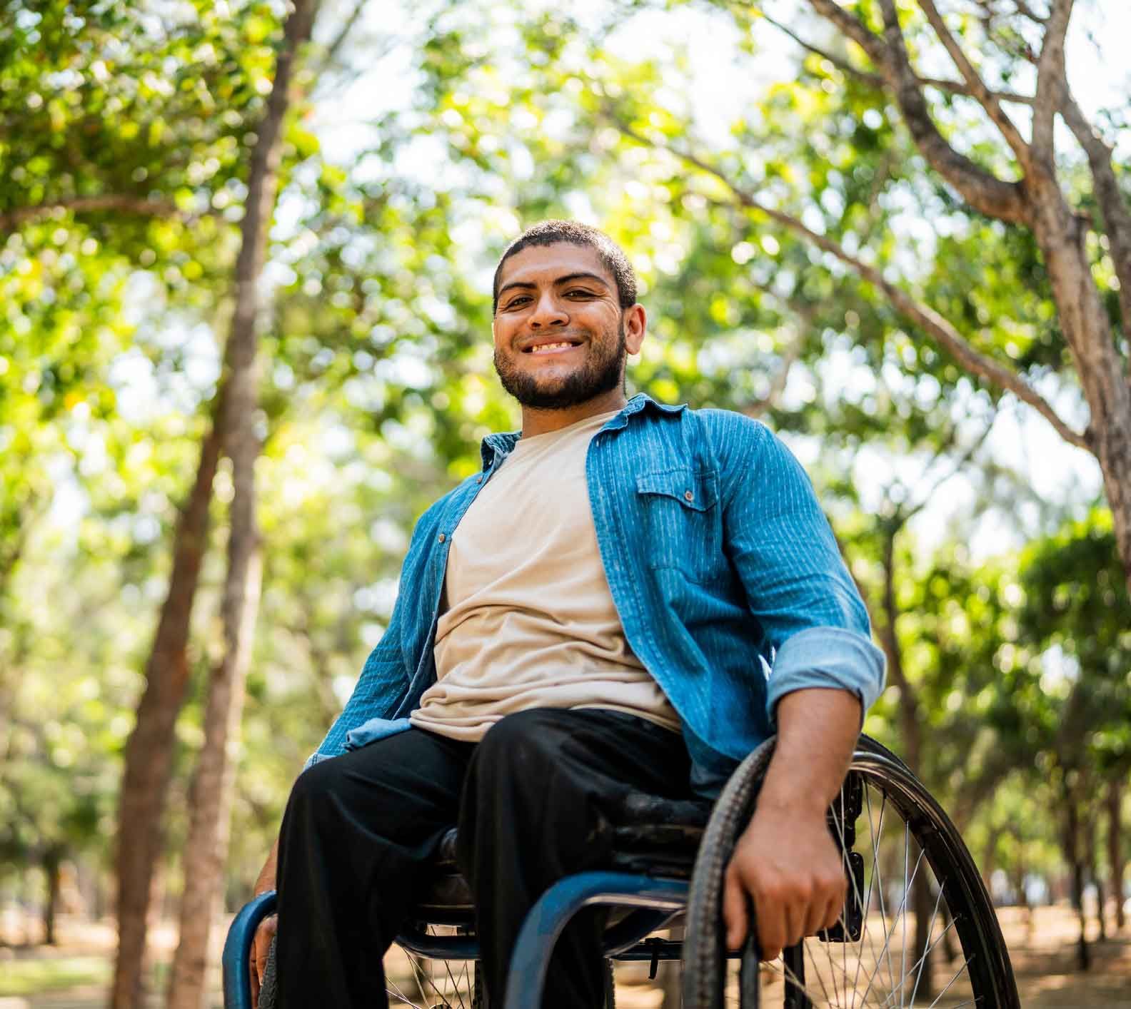 Smiling man in a wheelchair outdoors, wearing a blue shirt and tan shirt, with a forest in the background.