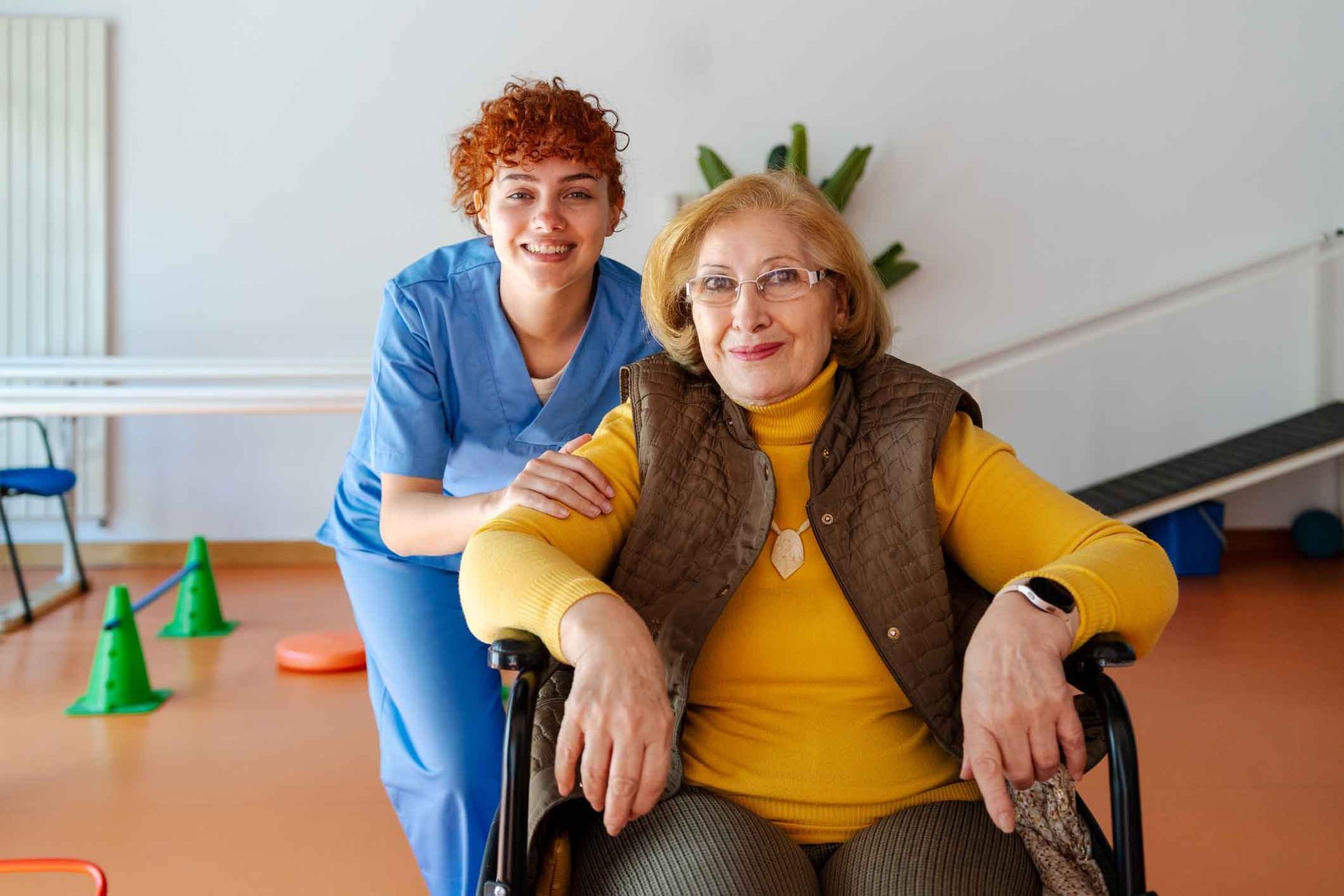 A smiling caregiver in blue scrubs assists a senior woman in a wheelchair wearing a yellow sweater and vest. They are in a therapy room.