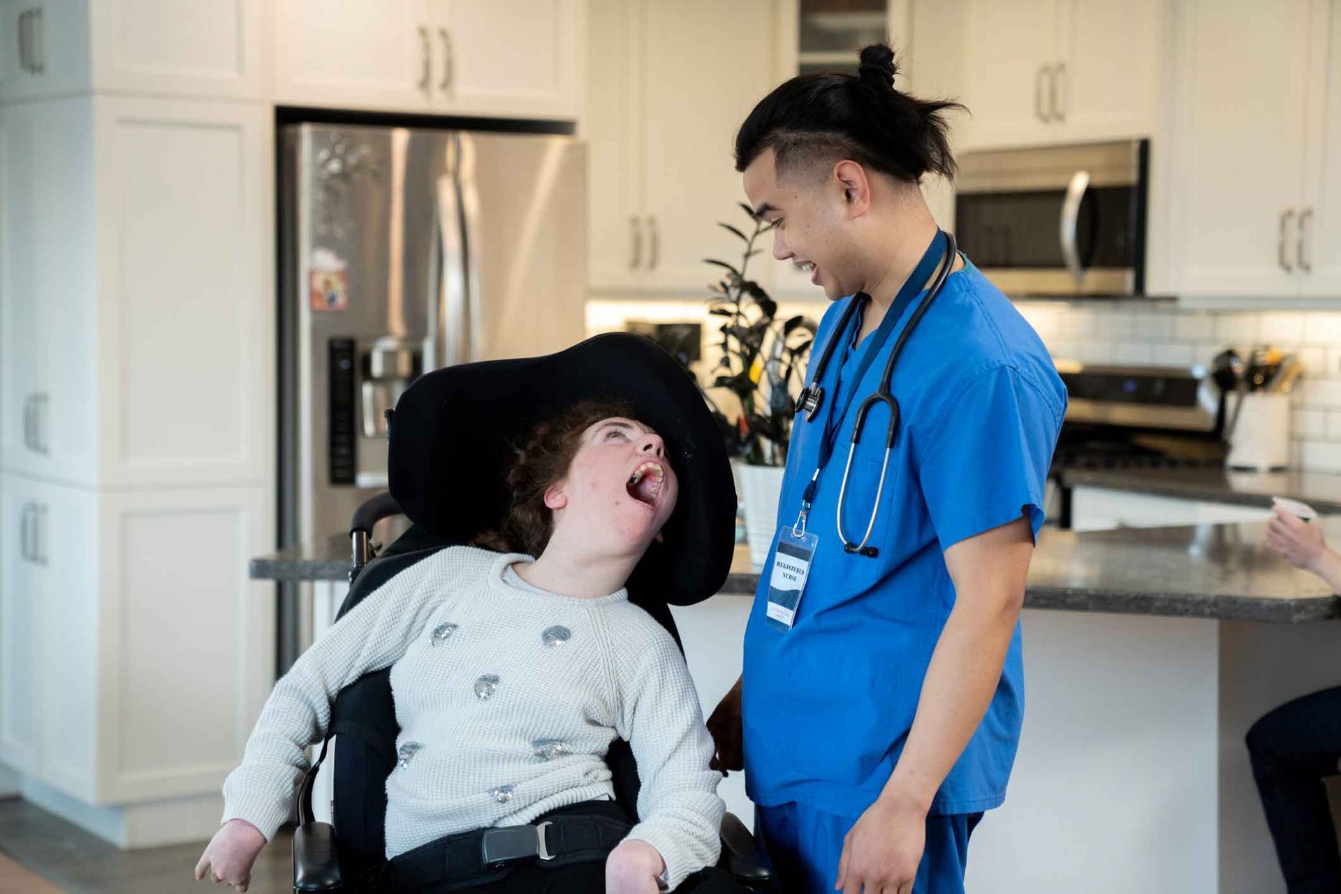 A young woman in a wheelchair with an open mouth looks up at a smiling healthcare worker. They are in a kitchen. A young woman in a wheelchair with an open mouth looks up at a smiling healthcare worker. They are in a kitchen.