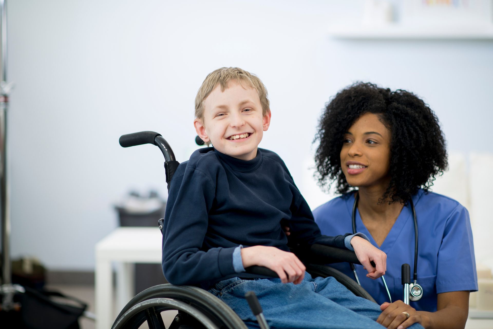 Healthcare worker kneels beside smiling child in wheelchair.