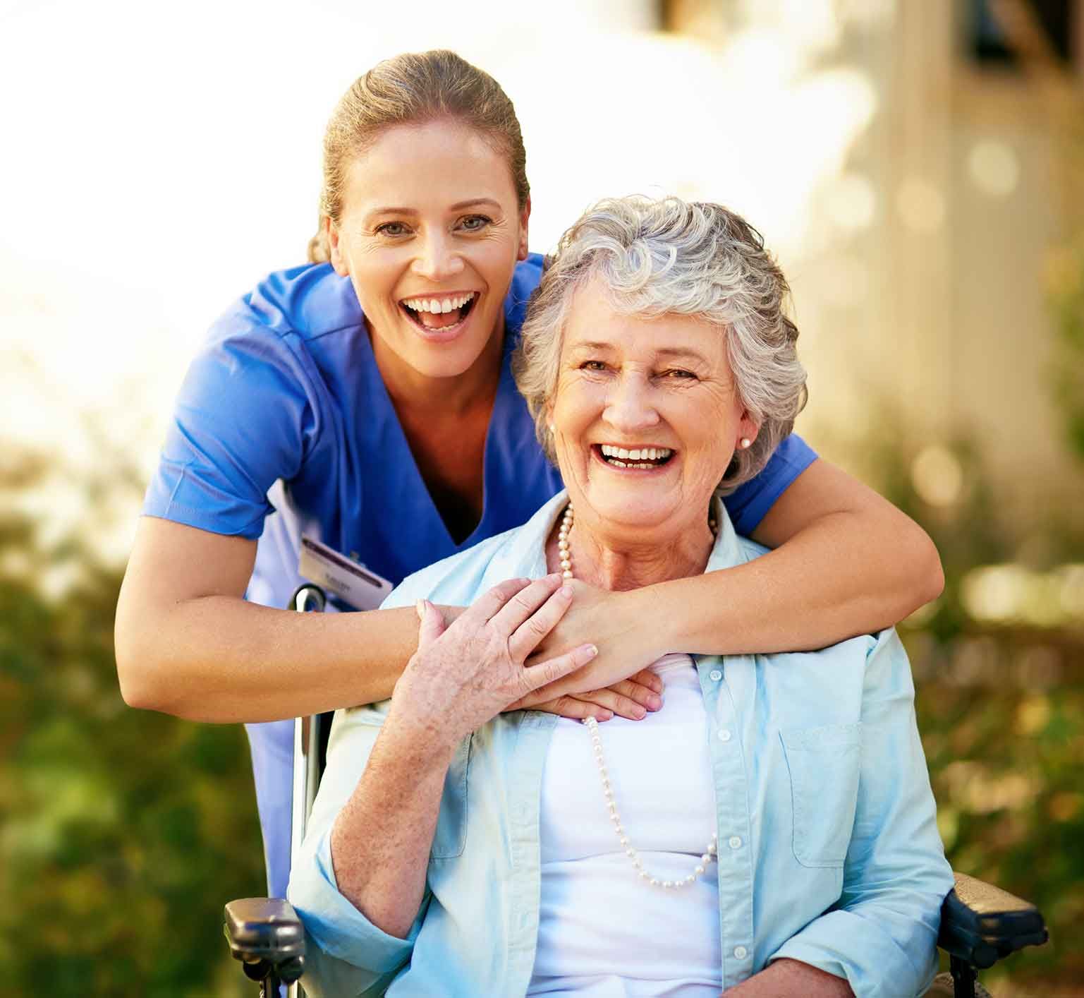Smiling young caregiver embraces an elderly woman in a wheelchair, both outdoors. They are both laughing.