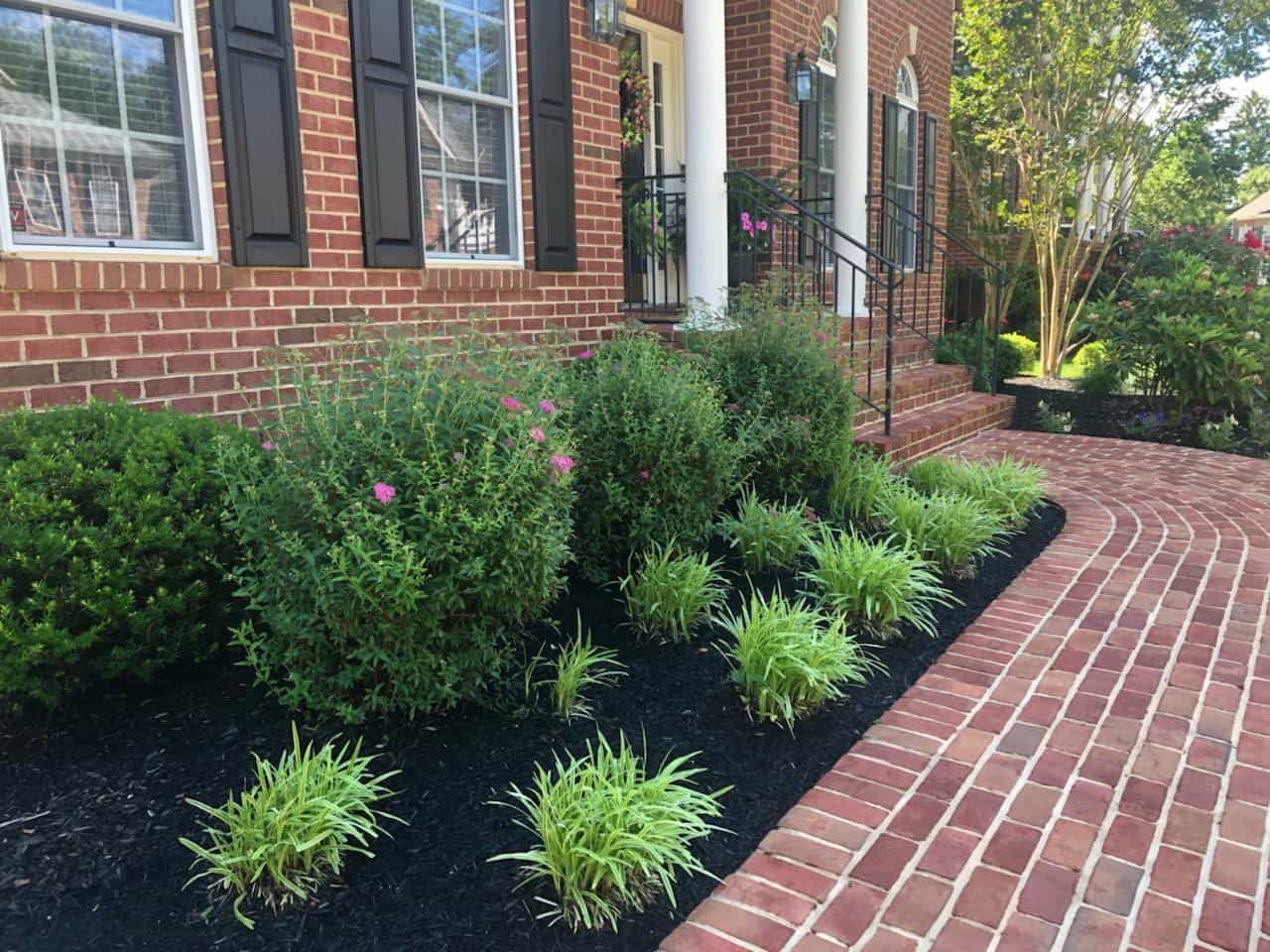 A brick walkway leading to a brick house with a lush green garden in front of it.