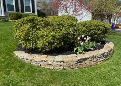 A stone planter with bushes and flowers in front of a house.