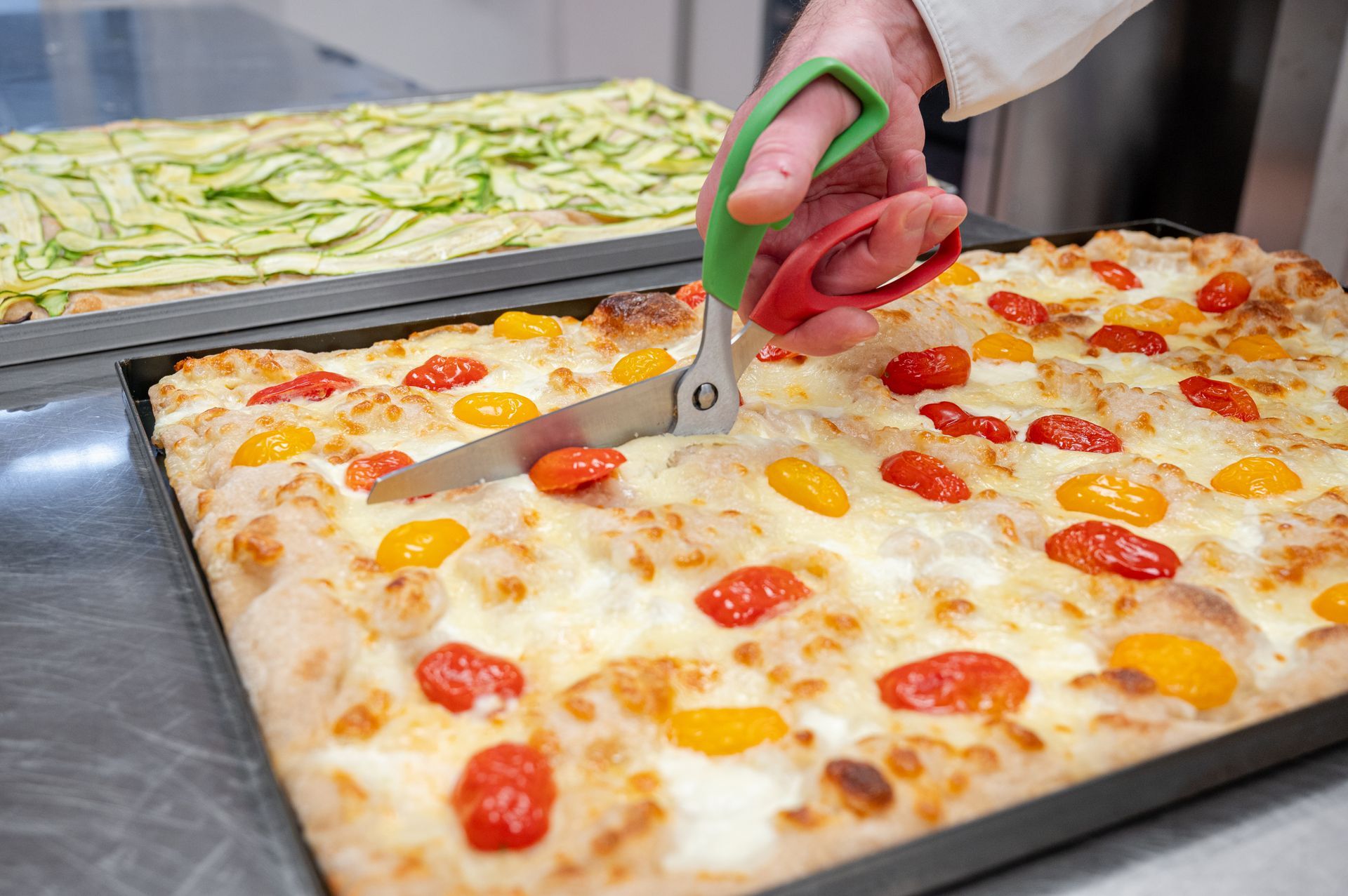 Una persona taglia una pizza teglia con una rotella tagliapizza dal manico verde; nelle vicinanze si stanno cuocendo pizze fresche.
