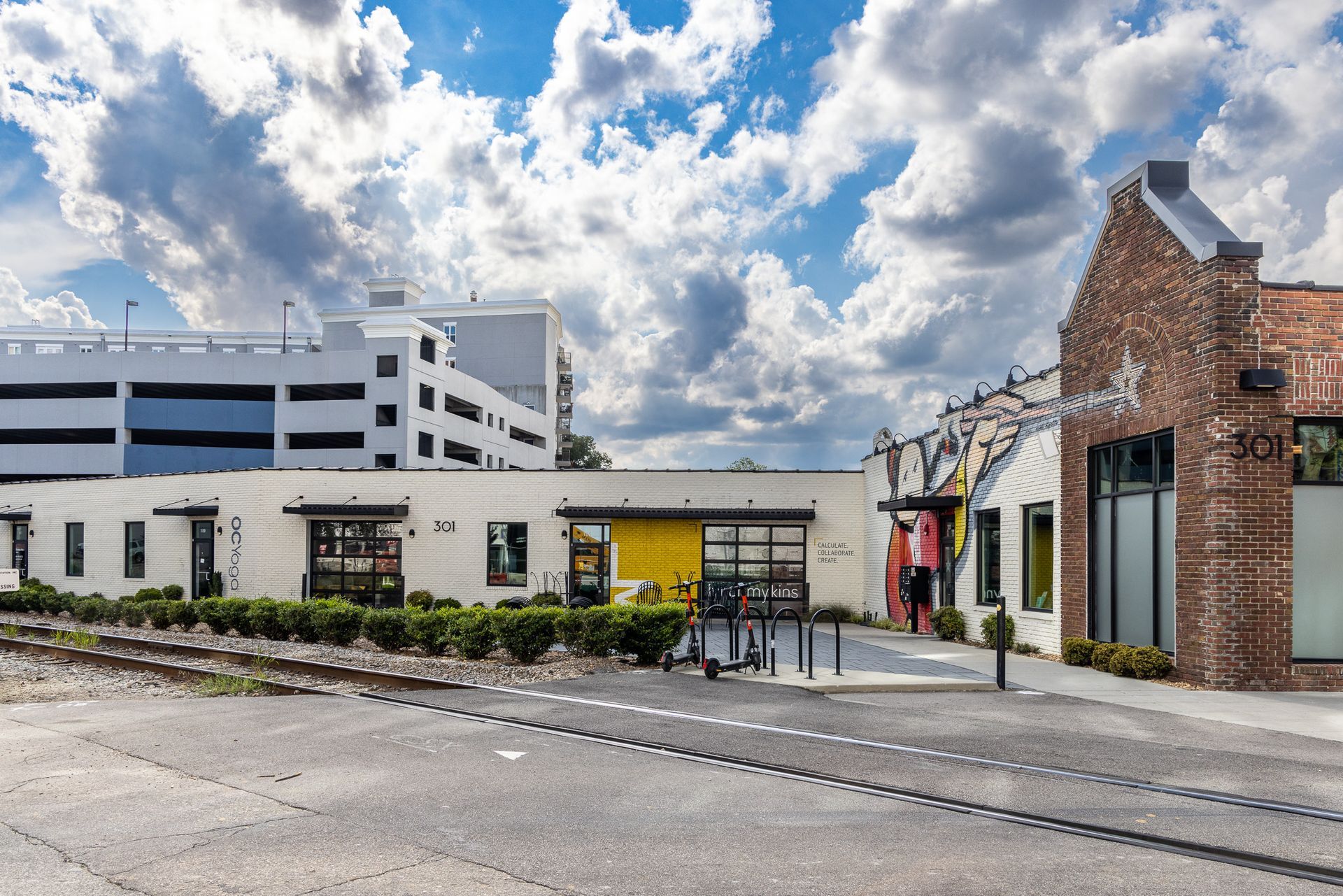 Buildings with a parking garage, brick facade, and train tracks under a cloudy sky.