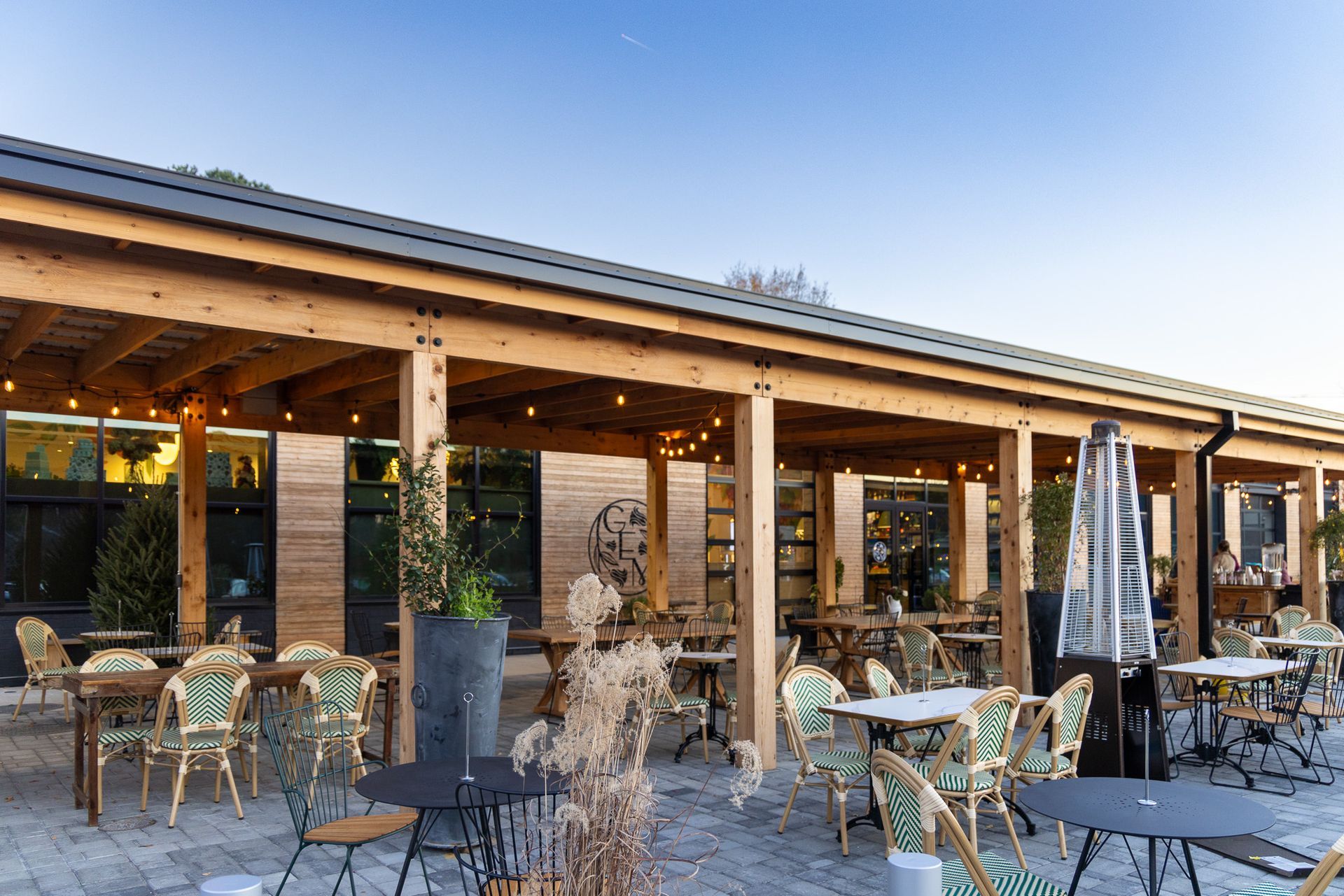 Outdoor restaurant patio with tables, chairs, and heaters under a wooden roof.