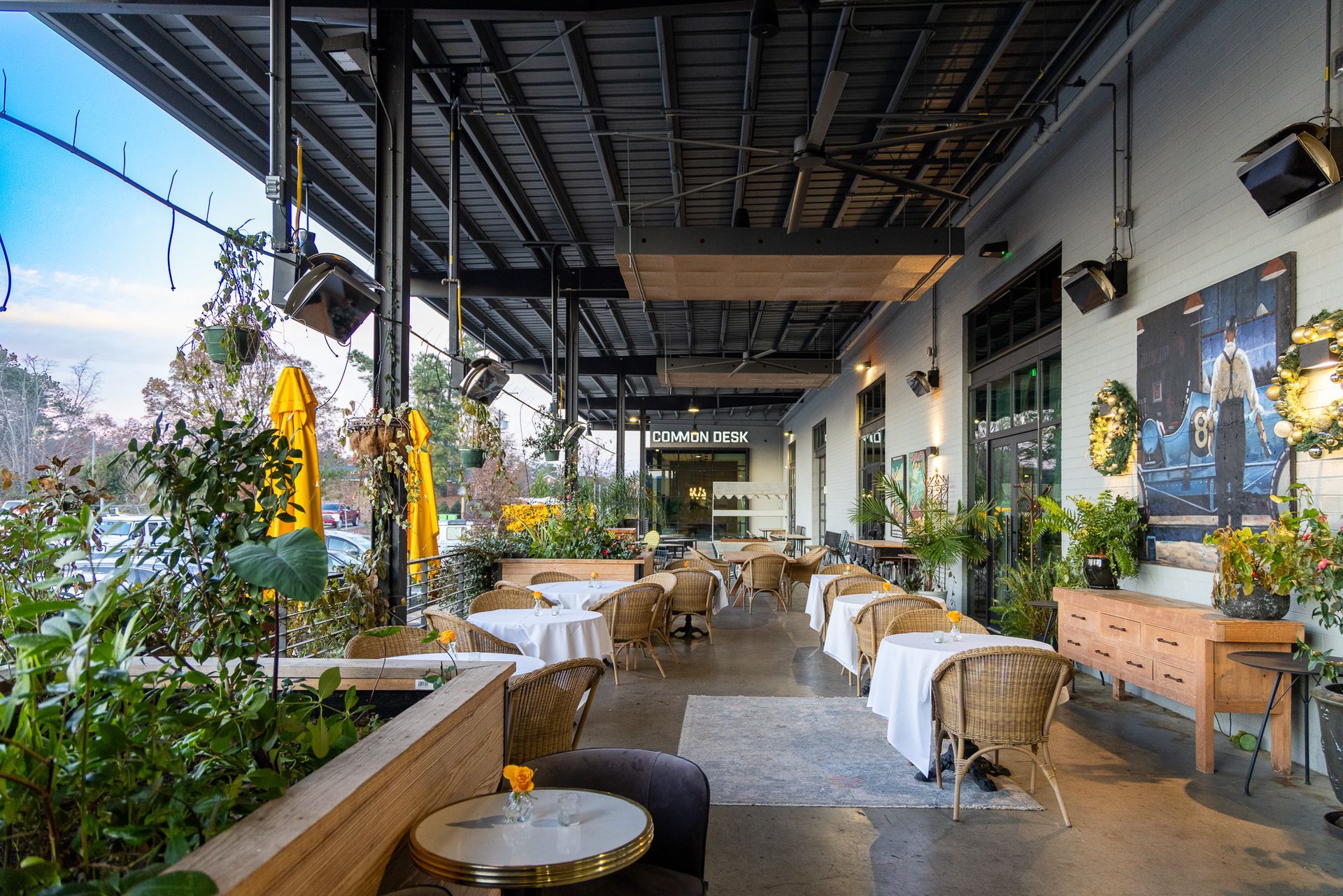 Outdoor restaurant patio with tables, chairs, plants, and a light-colored building.
