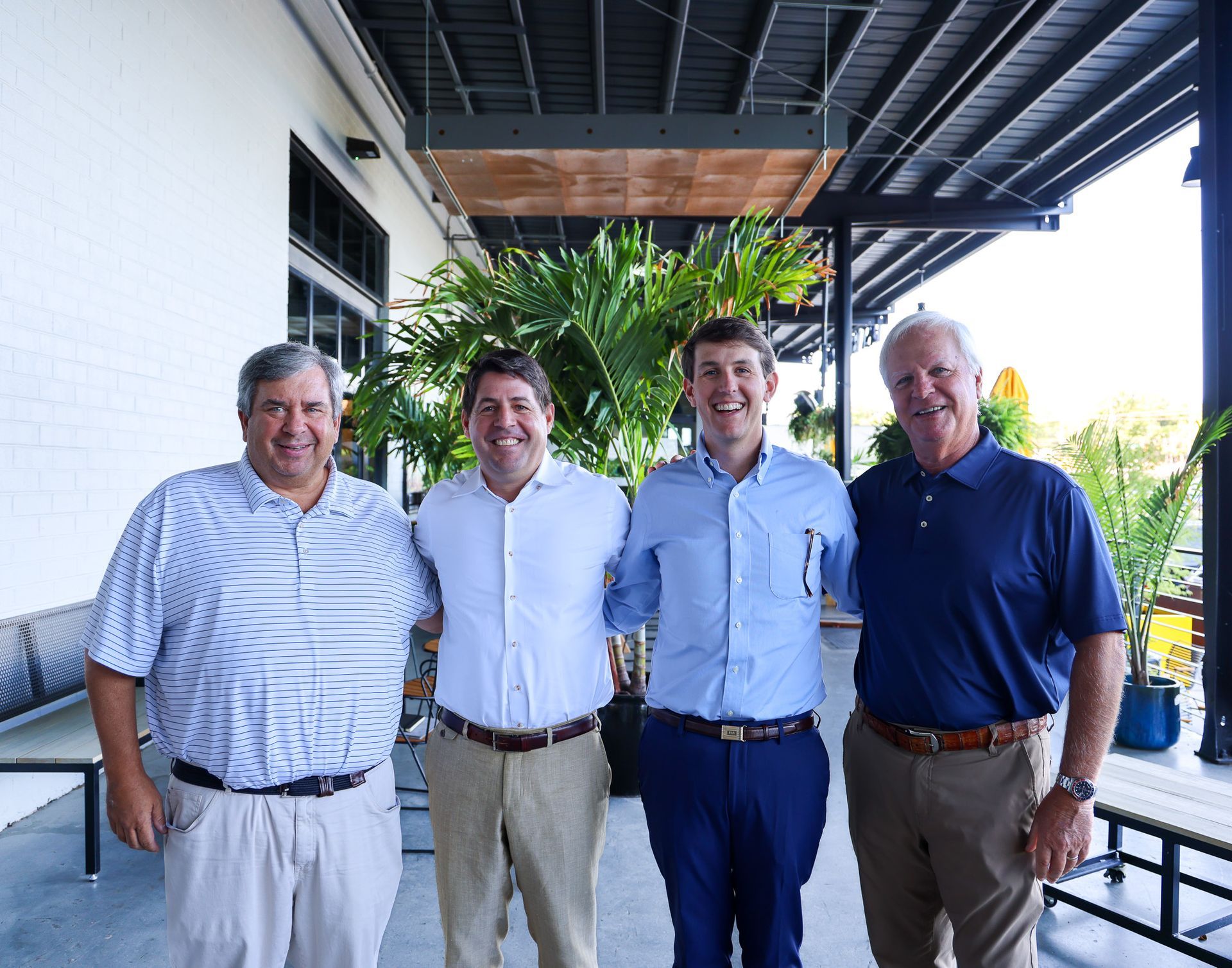 Four men standing outside. They wear shirts, khakis. One man is smiling.