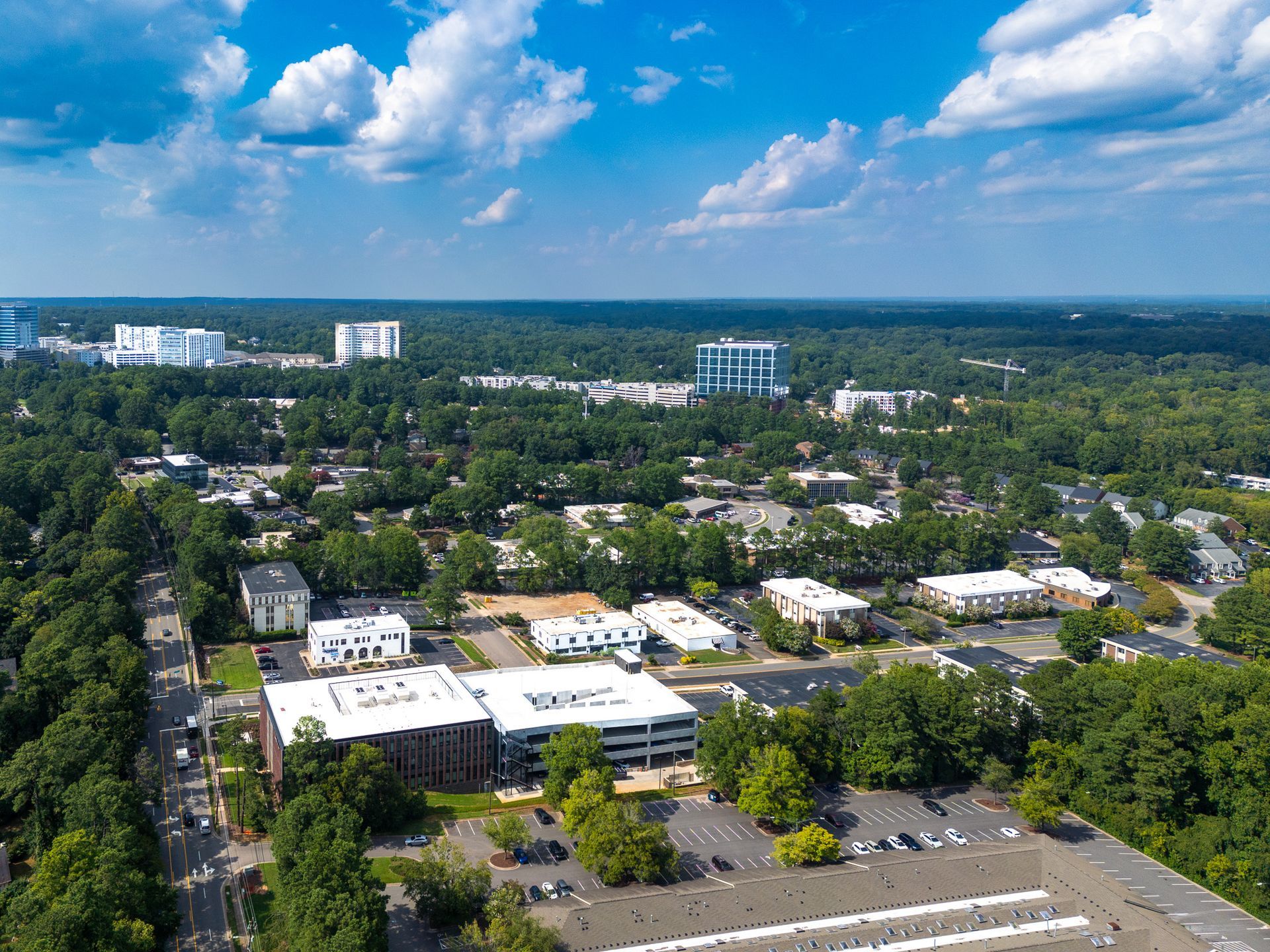 Aerial view of a suburban area with buildings, trees, a parking lot, and a blue sky with clouds.