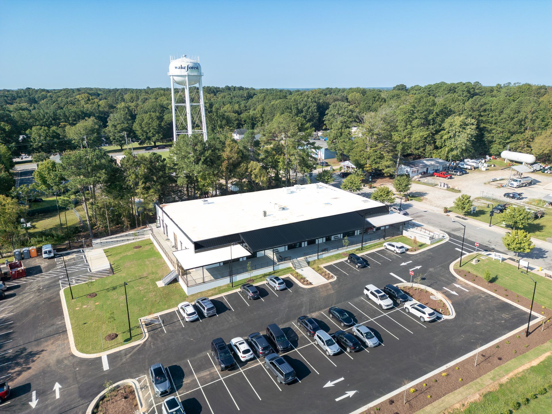 Aerial view of a modern building with a parking lot, water tower in background, and surrounding trees under a blue sky.