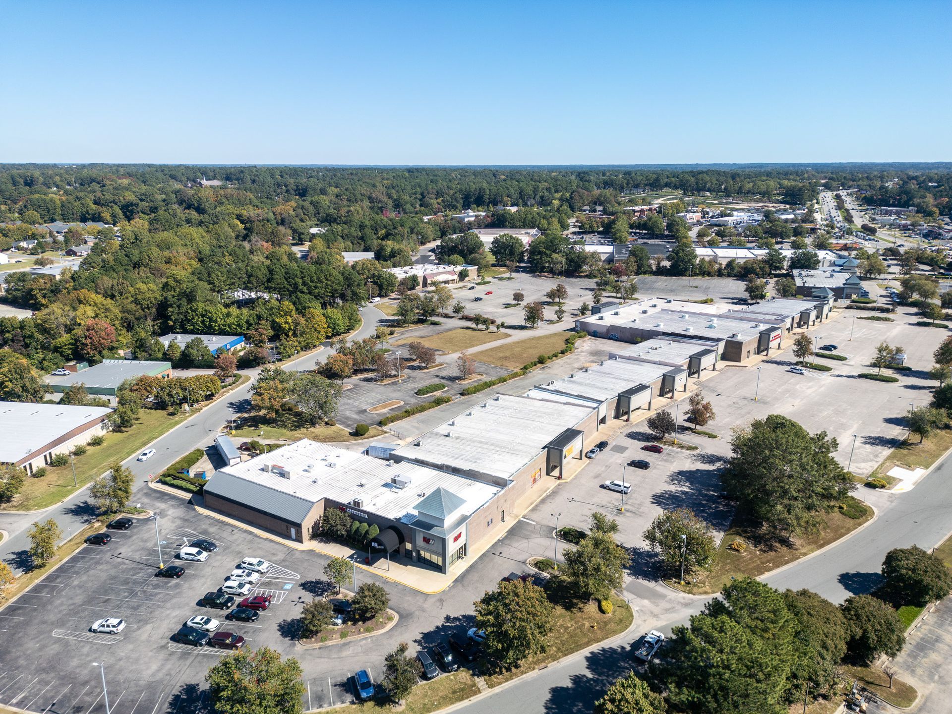 Aerial view of a mostly empty shopping center on a sunny day; parking lots, trees, buildings.
